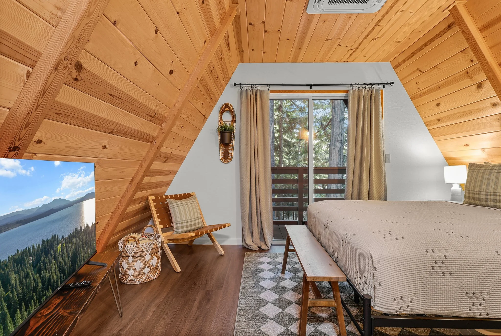Bedroom with wooden sloped ceiling, large window with beige curtains, a bed with a beige patterned blanket, a small wooden bench at the foot of the bed, a plaid pillow, a wooden chair with plaid cushion, a flat-screen TV showing a lake and mountain l