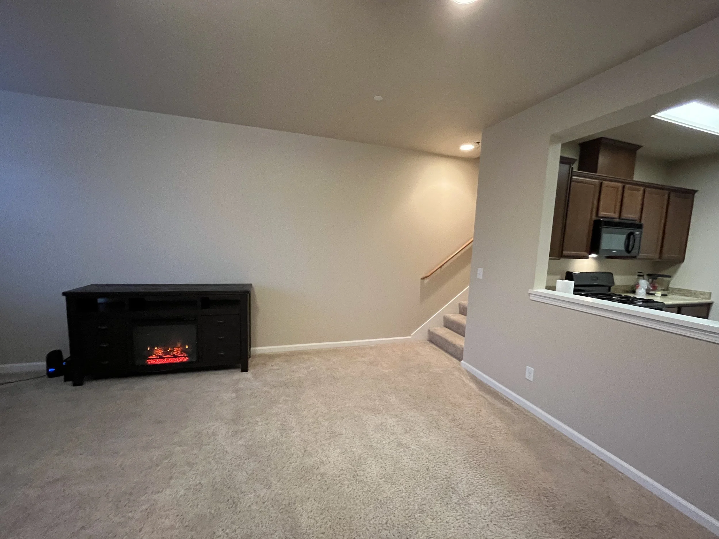 Living room with beige carpeting, a small electric fireplace, and kitchen in the background with wooden cabinets and a countertop.