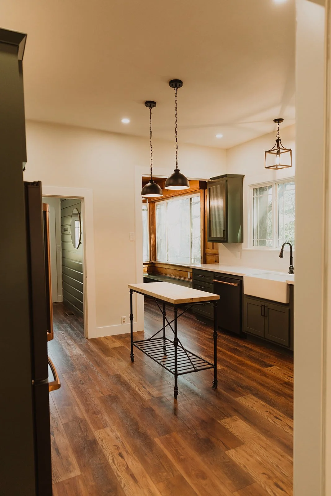 A kitchen with wooden flooring, black cabinets, and a farmhouse sink. There are three pendant lights hanging from the ceiling and a small table with a marble top and metal legs in the center.