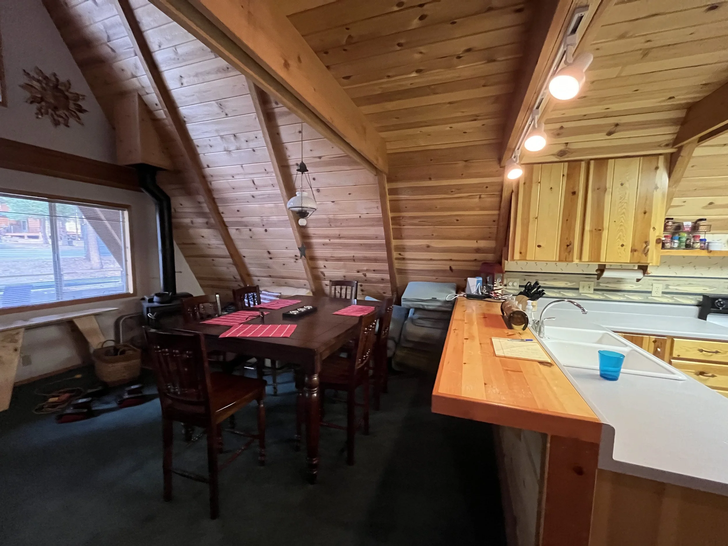 Interior view of a wooden cabin kitchen and dining area with a sloped wood-paneled ceiling, a wooden dining table set with red placemats, and a window with blinds showing an outdoor deck.