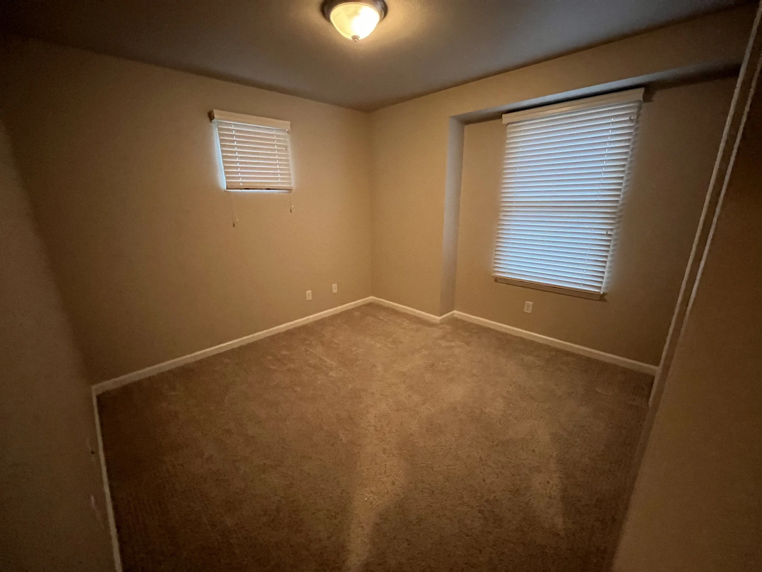 Empty room with beige walls, two windows with blinds, and a ceiling light fixture.