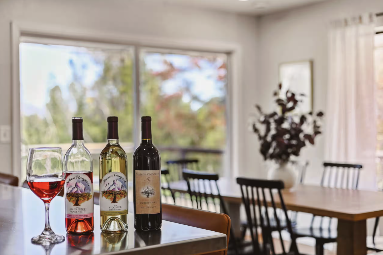 Three bottles of wine, a wine glass filled with red wine, and a wine cork on a kitchen island, with a dining area and large windows showing an outdoor view in the background.