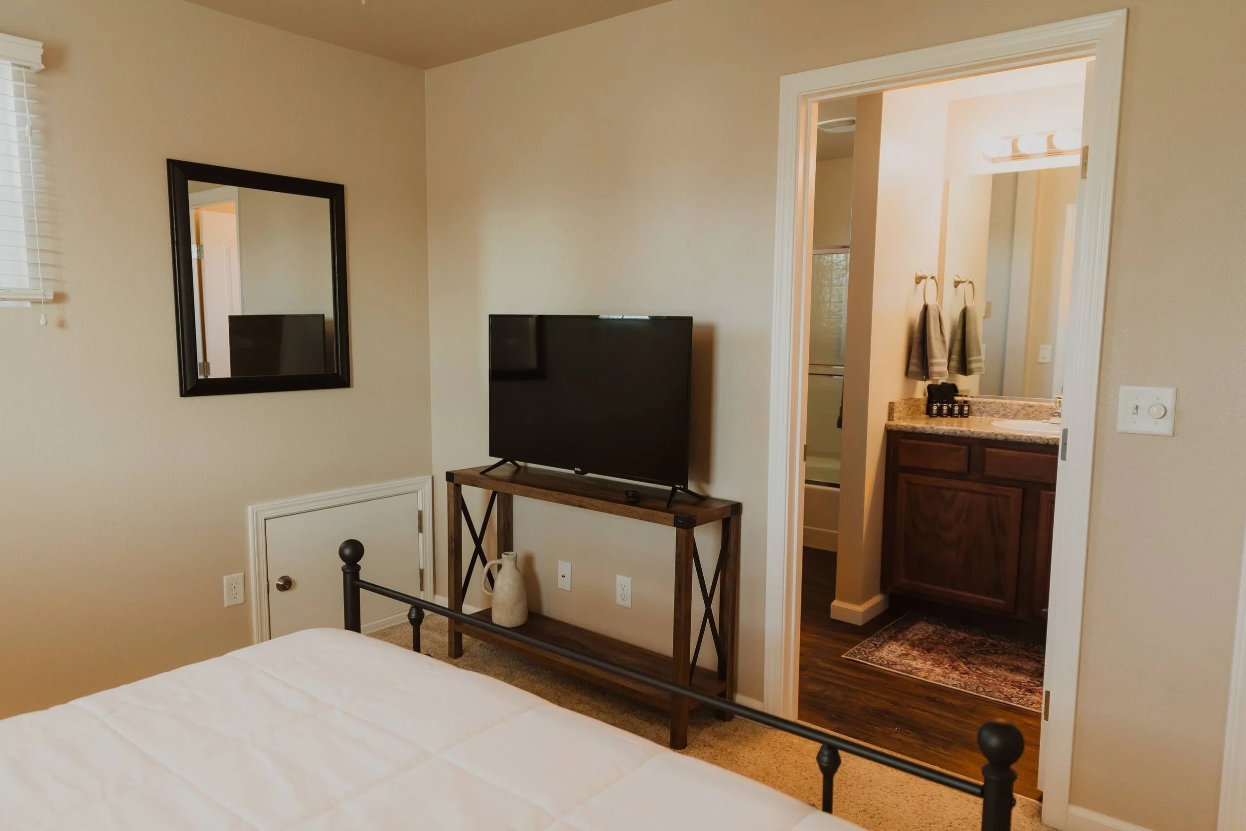 A bedroom with beige walls, a black-framed mirror on the wall, a flat-screen TV on a wooden table, a vase on the floor, and a doorway leading to a bathroom with a vanity, mirror, and bathroom towels.