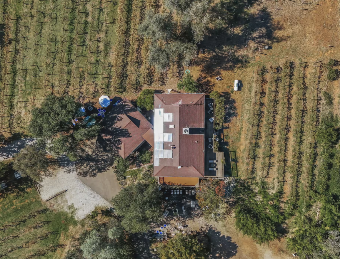 Aerial view of a house surrounded by trees and open land, with a garden and outdoor seating area.
