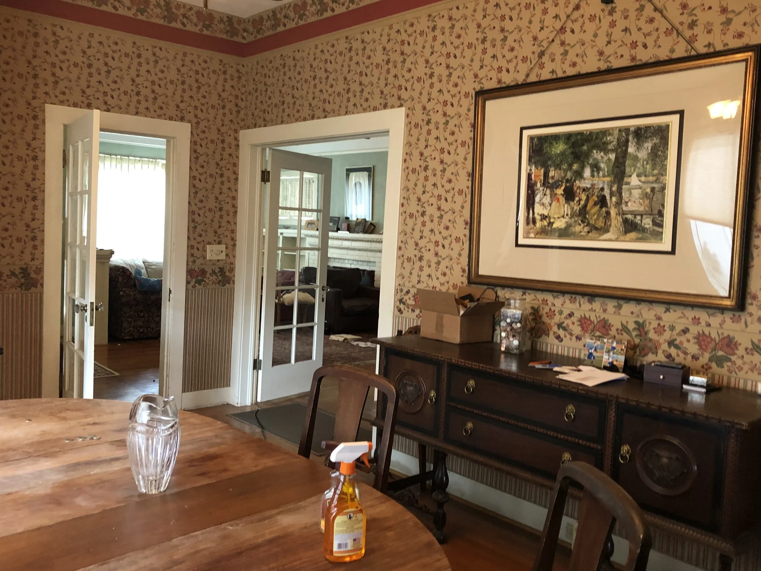 View of a dining room with floral wallpaper, a wooden table with a glass of water and cleaning spray, and a sideboard with papers. There are open French doors leading to other rooms with windows, chairs, and pictures visible.