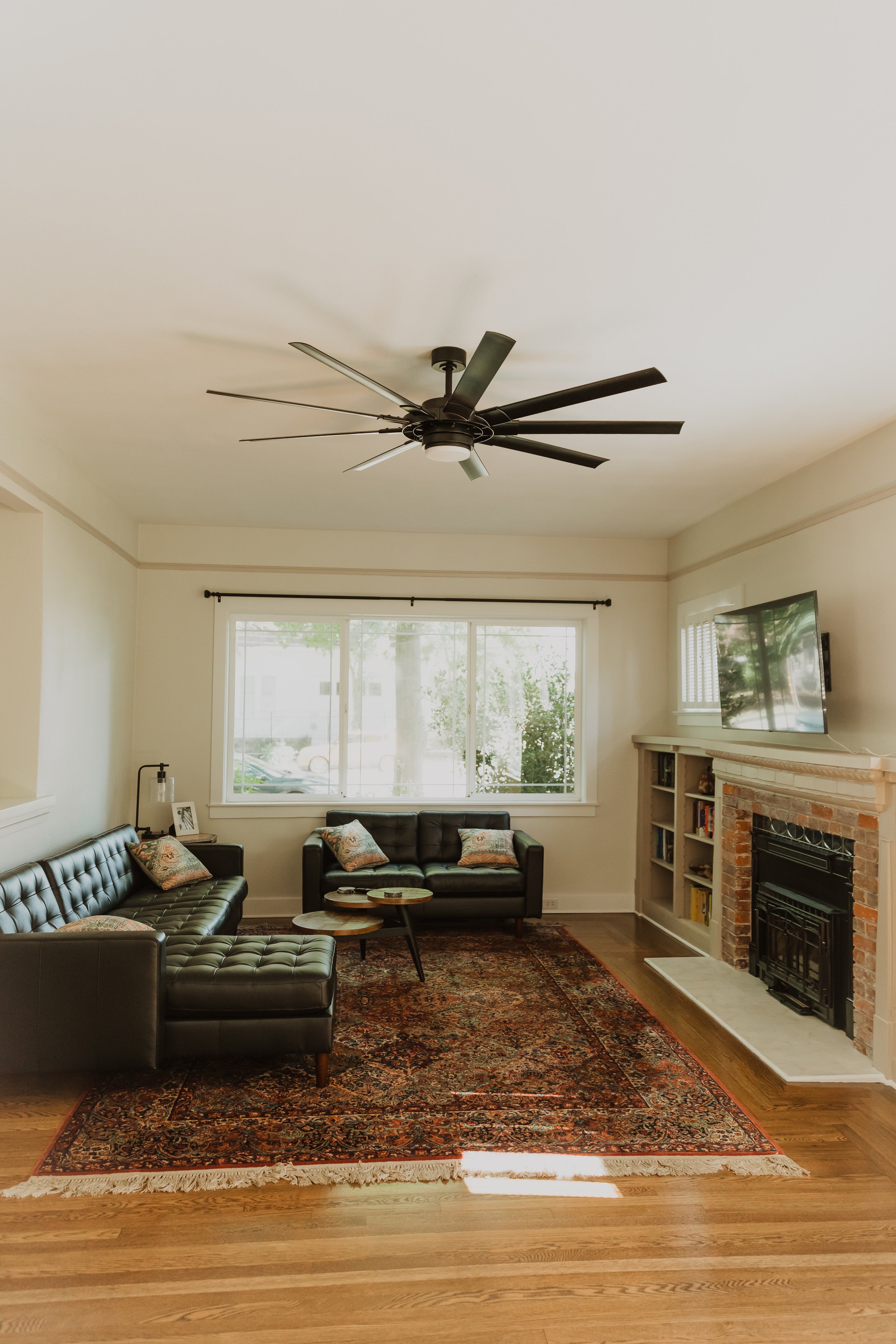 Living room with black leather sectional sofa, wooden coffee tables, patterned area rug, brick fireplace, television, window with blinds, and ceiling fan.