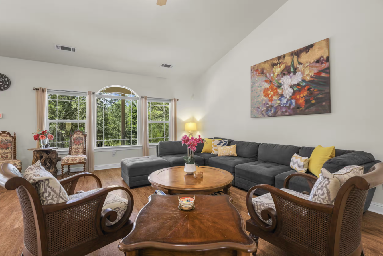 Living room with a large gray sectional sofa, wooden coffee table, vintage armchairs, colorful cushions, floral artwork on the wall, window with trees outside, and decorative items on the tables.