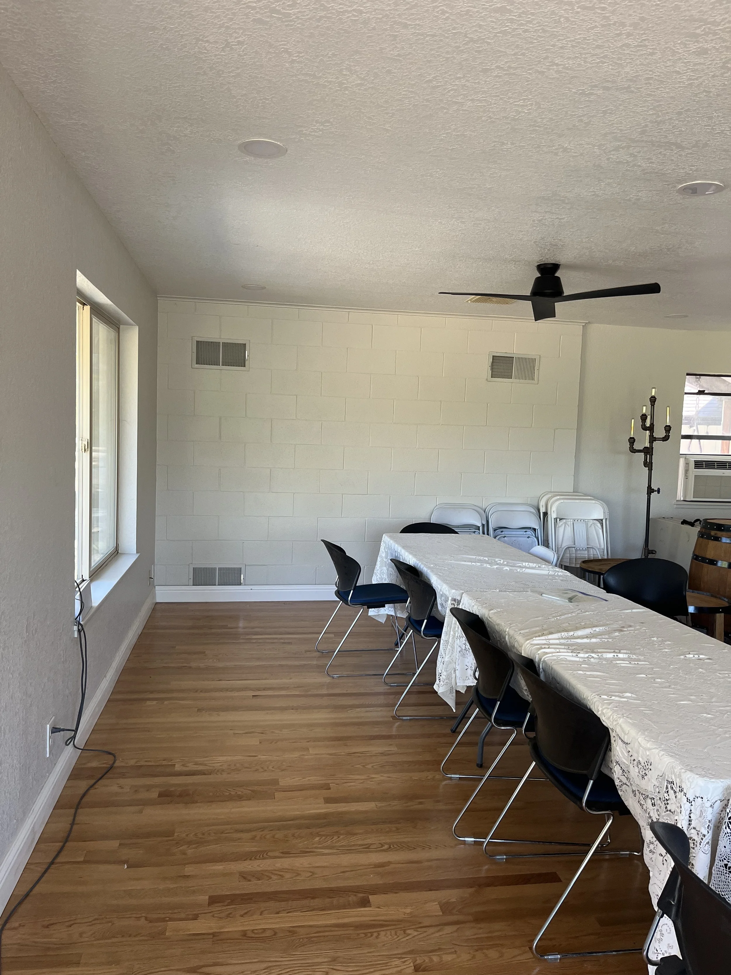 A long table with a white lace tablecloth and black chairs around it in a room with wooden floor and white walls. There are several stacked white chairs and a candelabra against the wall, windows letting in natural light, and a black ceiling fan.