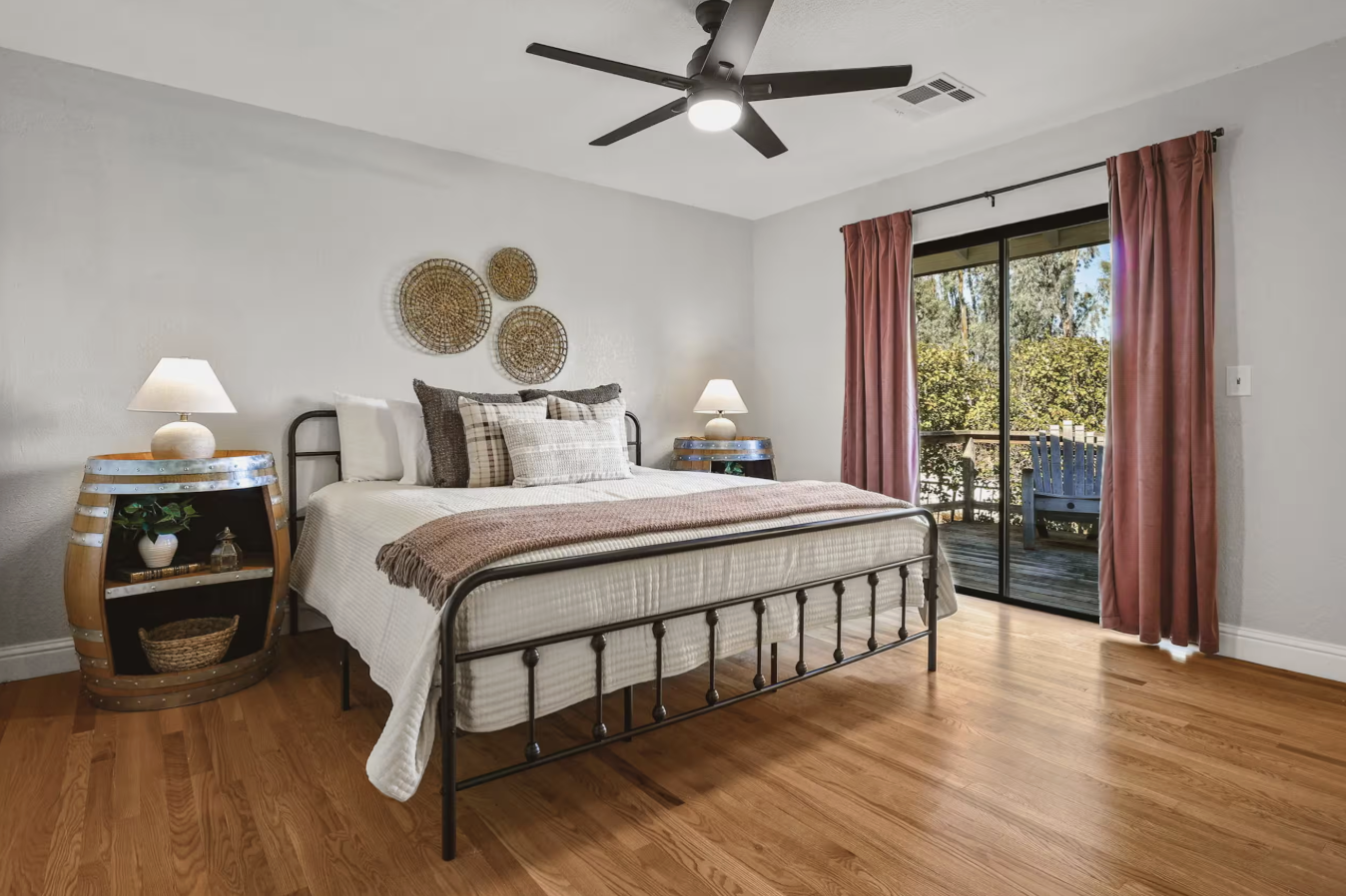 Bedroom with a metal bed frame, white bedding, brown and beige pillows, two wooden nightstands with lamps, pink curtains, wooden floor, and a sliding glass door leading to a balcony with outdoor furniture.