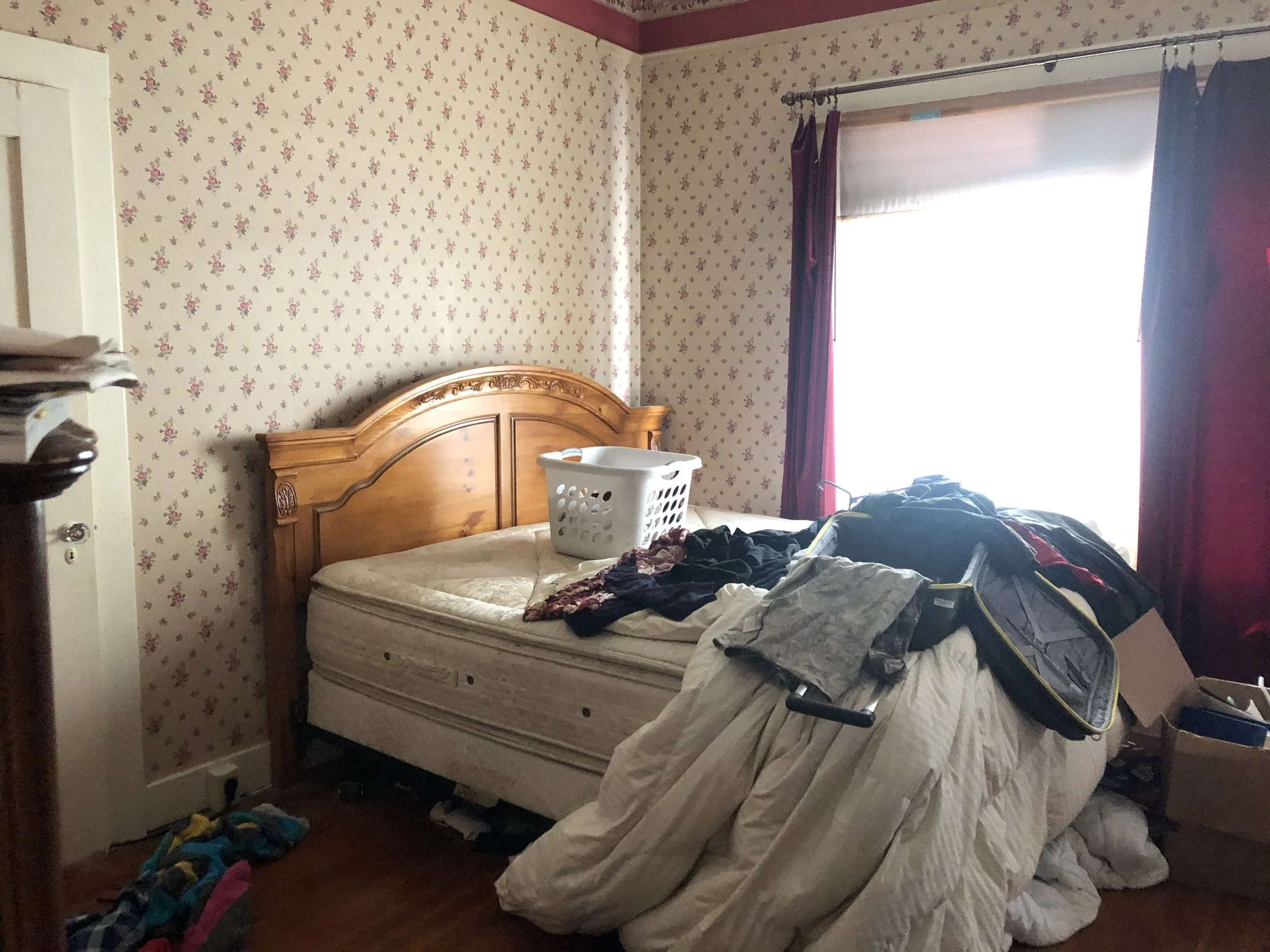 A cluttered bedroom with two beds, one with a mattress and pillow, and clothing scattered on the beds and floor. There is a laundry basket on the bed and a window with purple curtains letting in light. The wallpaper has a floral pattern, and a wooden