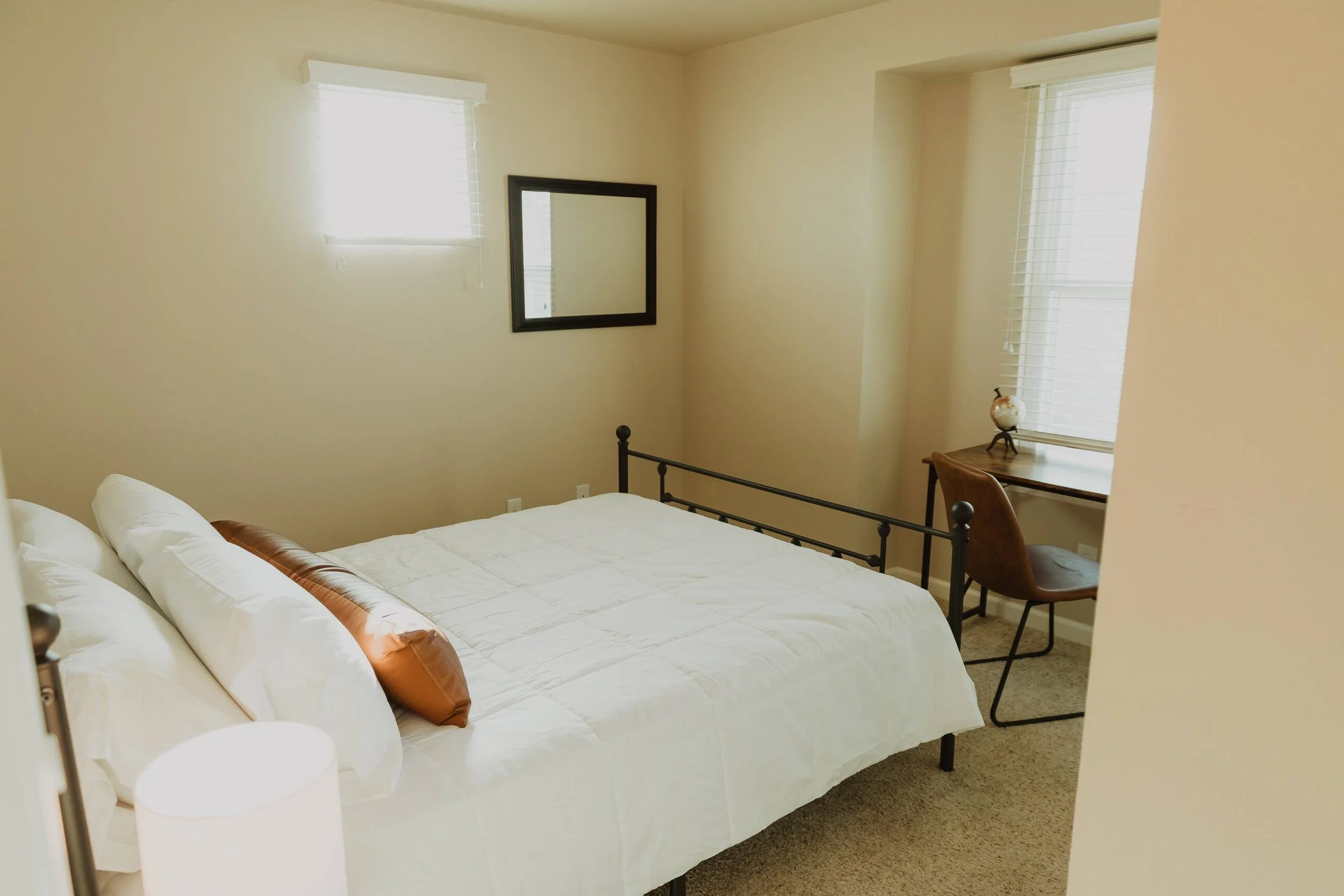A bedroom with an unmade bed, two windows with blinds, a mirror on the wall, a small desk with a chair, and a decorative globe on the desk.