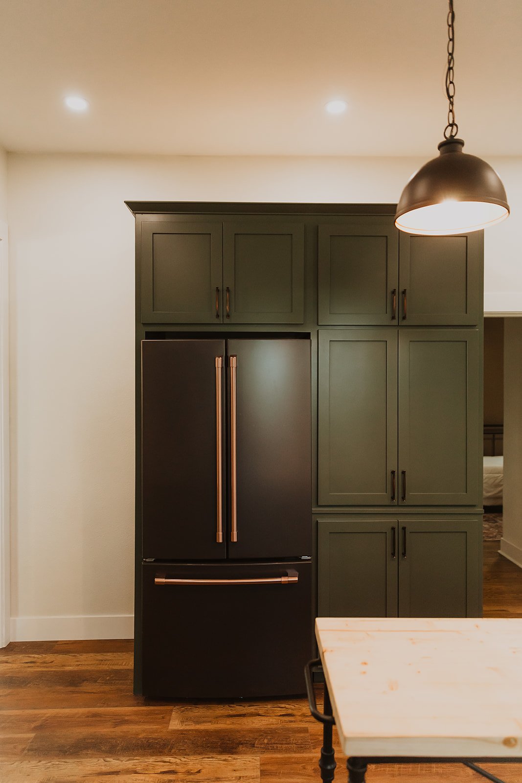 Kitchen corner with green cabinetry, black refrigerator with copper handles, and a pendant light overhead.