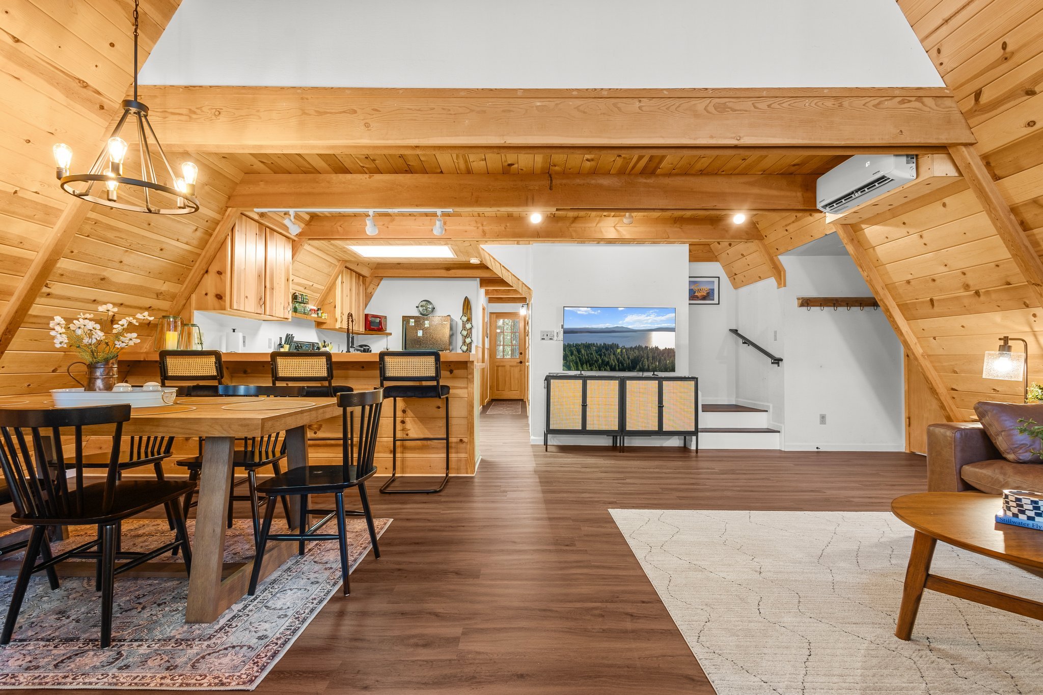 Interior of a living space with wooden walls and ceiling, a dining table with black chairs, a kitchen with a refrigerator and shelves, a television showing a scenic view, and a large window with a view of a body of water and trees.
