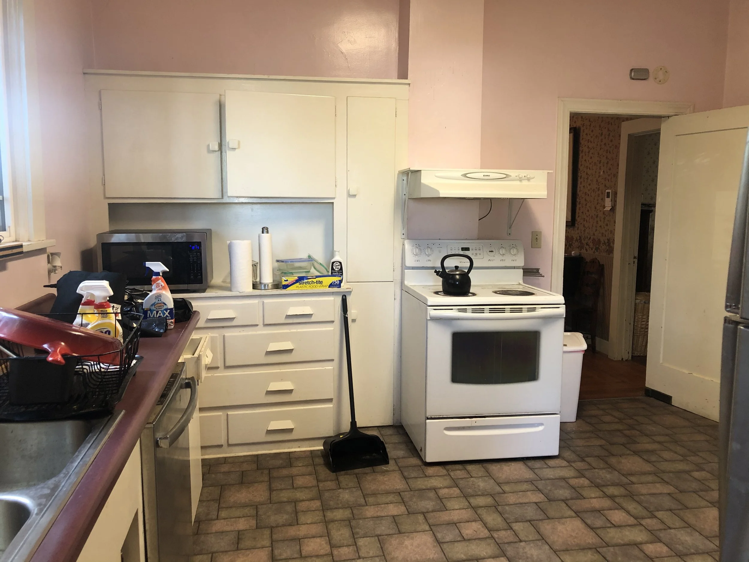 A small kitchen with pink walls featuring white cabinets, a white stove with a black teapot, a microwave, and a counter with cleaning supplies and paper towels.