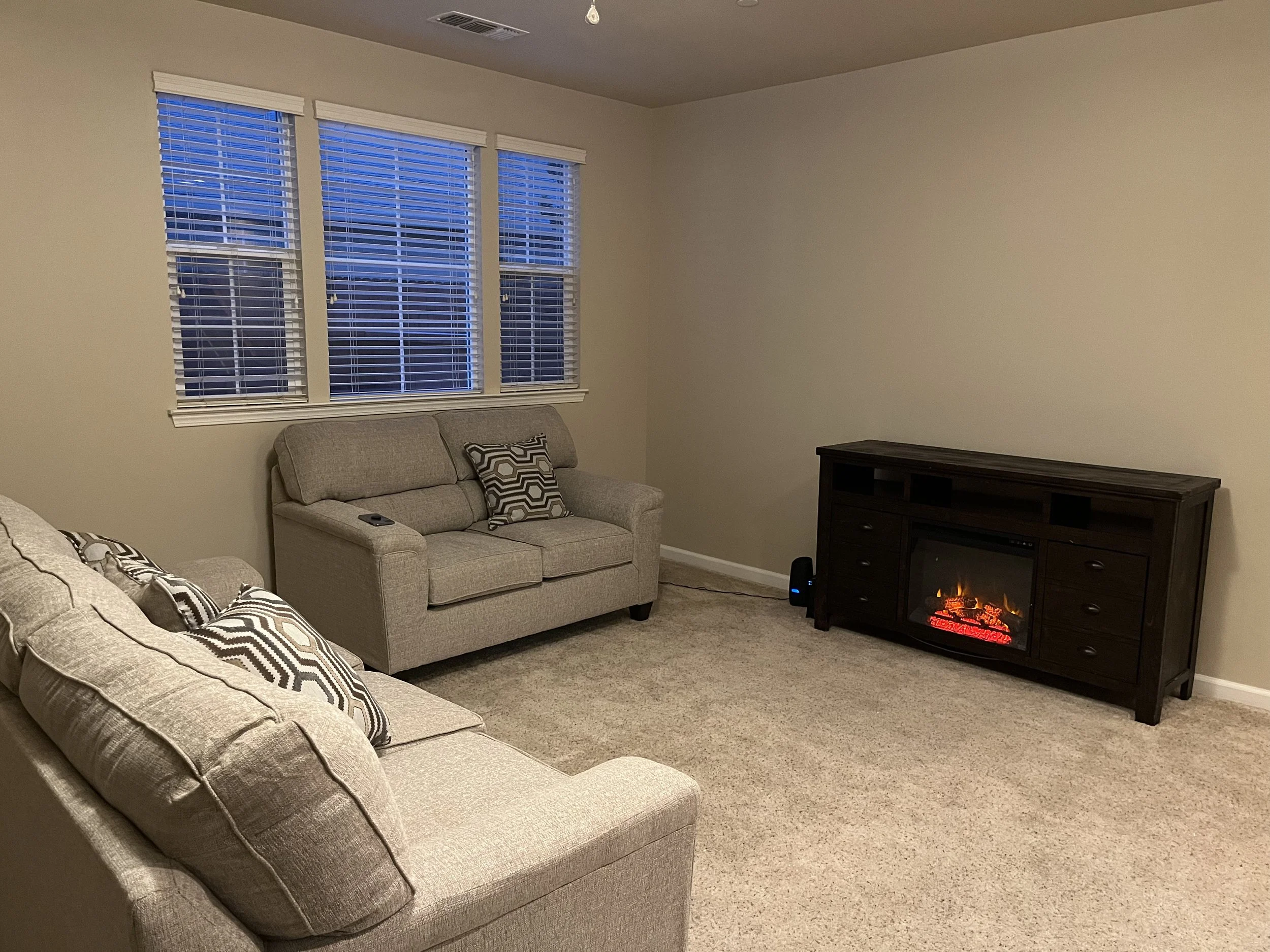Living room with beige sofas, decorative pillows, a dark wood entertainment center, a fireplace with a glowing fire effect, three windows with white blinds, and beige carpeting.