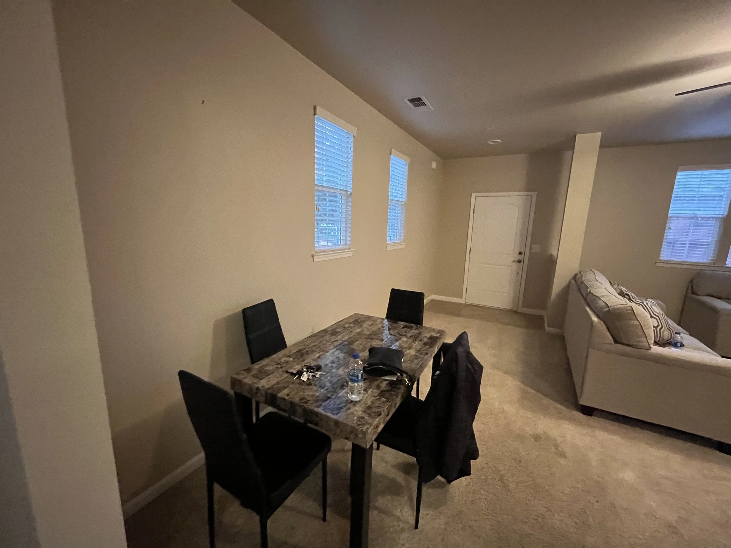 Living room with beige walls and multiple windows with blinds, featuring a dining table with chairs, water bottles, and some personal items, with a sofa and pillows in the background.