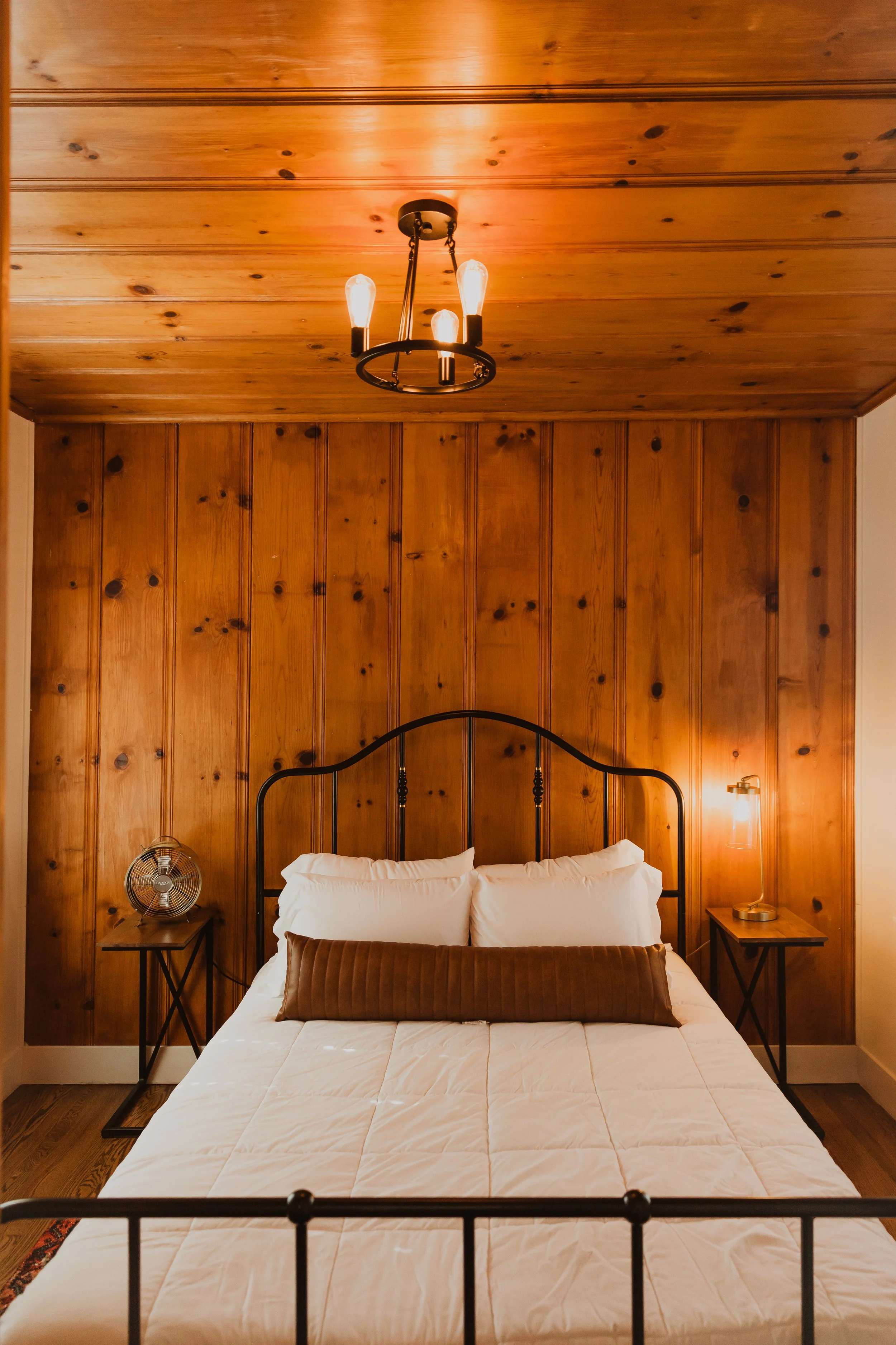 A bedroom with wood-paneled walls and ceiling, metal bed frame, white bedding, pillow, brown bolster pillow, wooden nightstands on each side, a round fan on the left nightstand, a brass table lamp on the right nightstand, and a ceiling chandelier wit