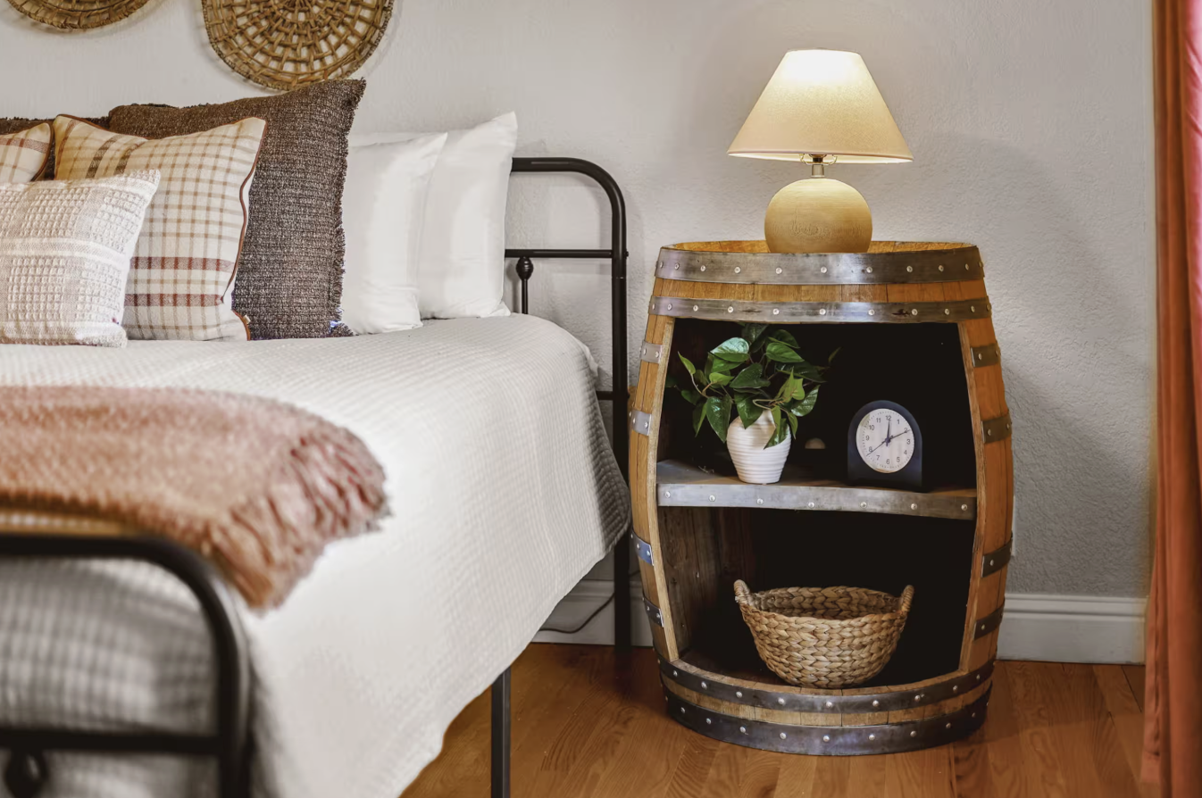 A bedroom corner with a bed and rustic barrel-shaped nightstand holding a lamp, plant, clock, and basket.