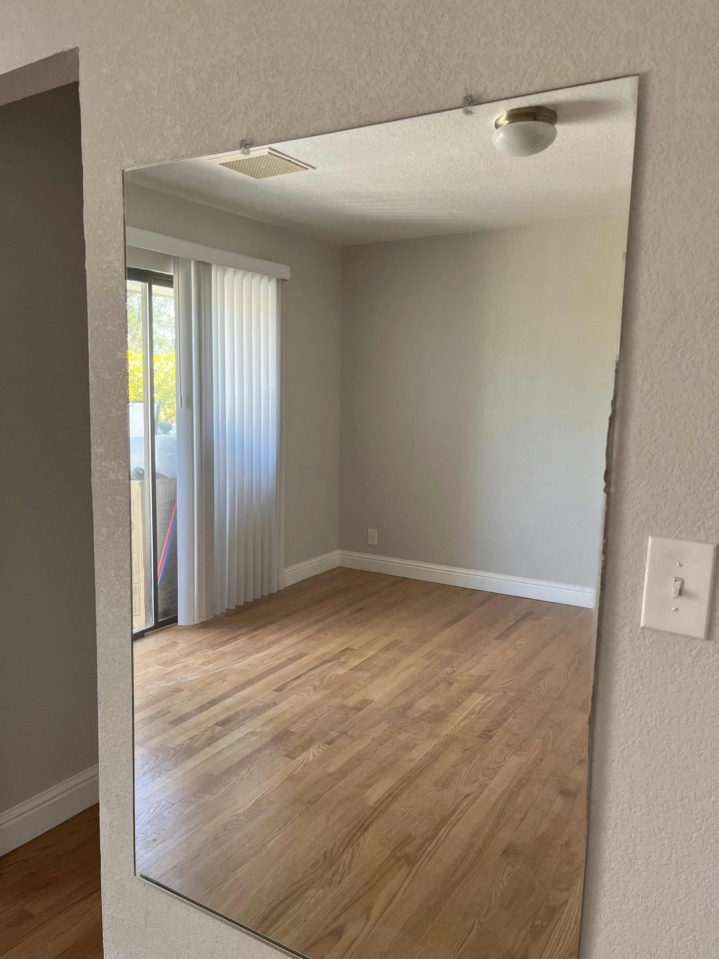 Empty living room corner with wooden floor, sliding glass door with vertical blinds, and a wall-mounted mirror reflecting the room.