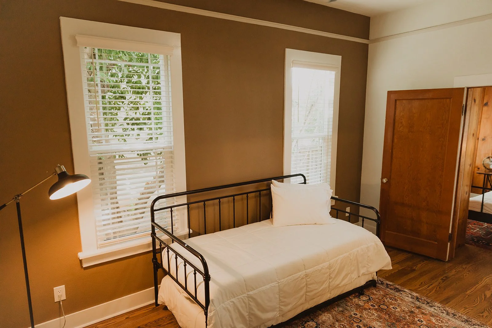 A bedroom with two windows with white blinds, a black metal bed with white bedding and a pillow, a wooden wardrobe, a floor lamp, and a patterned area rug.