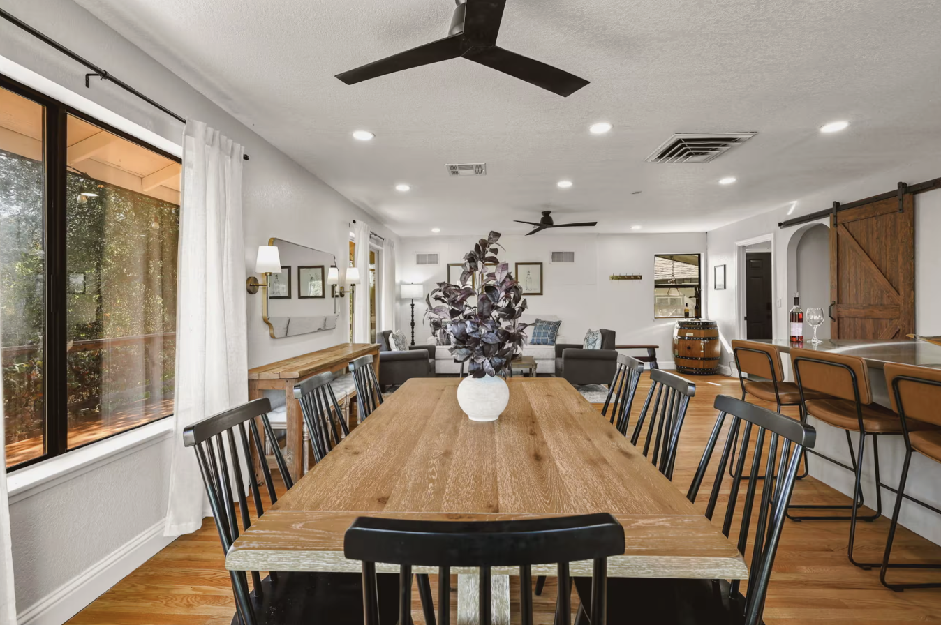 Open-concept dining and living room with a wooden dining table, black chairs, a large houseplant, a gray sofa, bar seating, and rustic decor.