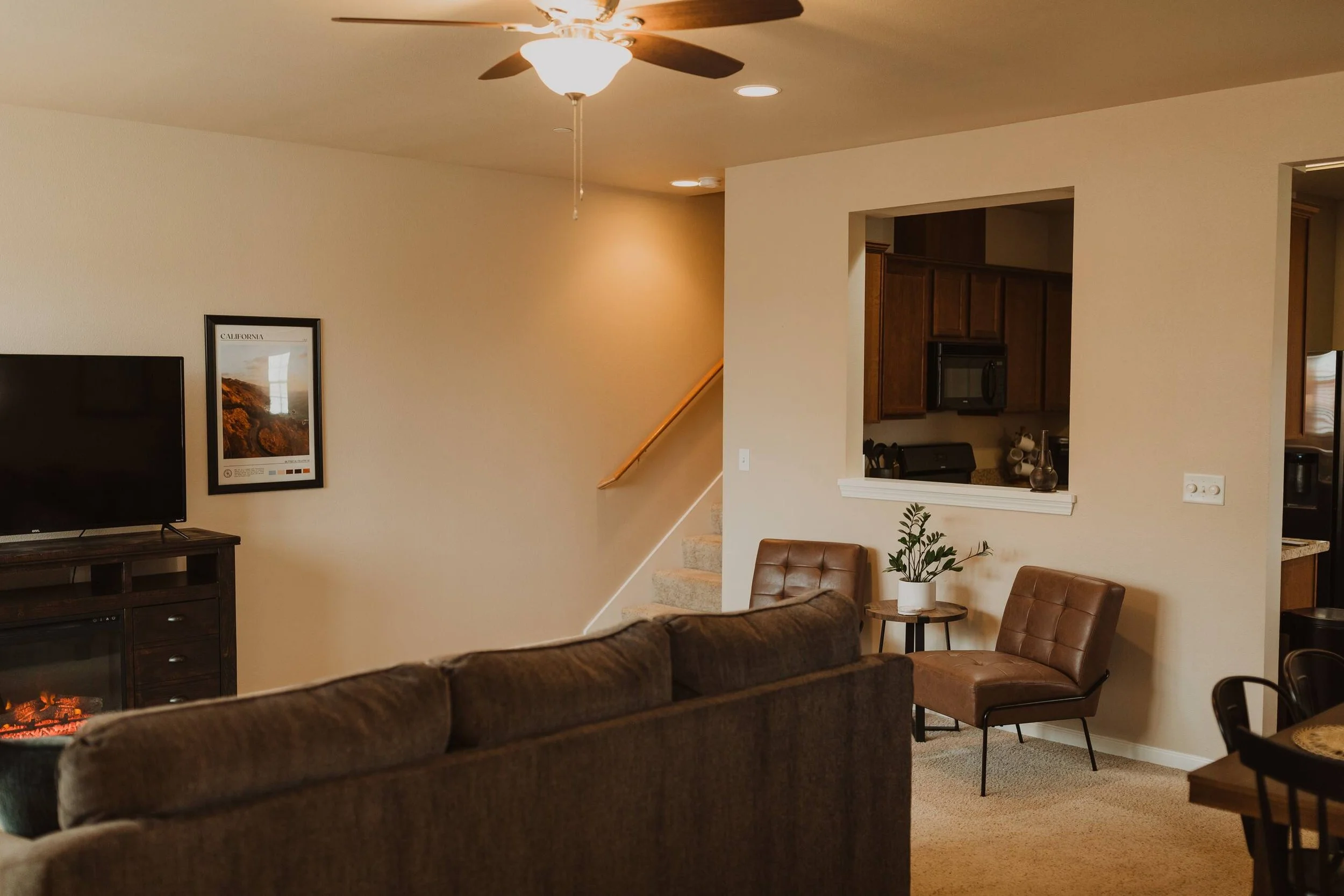 Living room with a ceiling fan, a wall-mounted TV, a framed California poster, a brown sofa, a brown leather chair, a small side table with a plant, and an open kitchen with wooden cabinets.