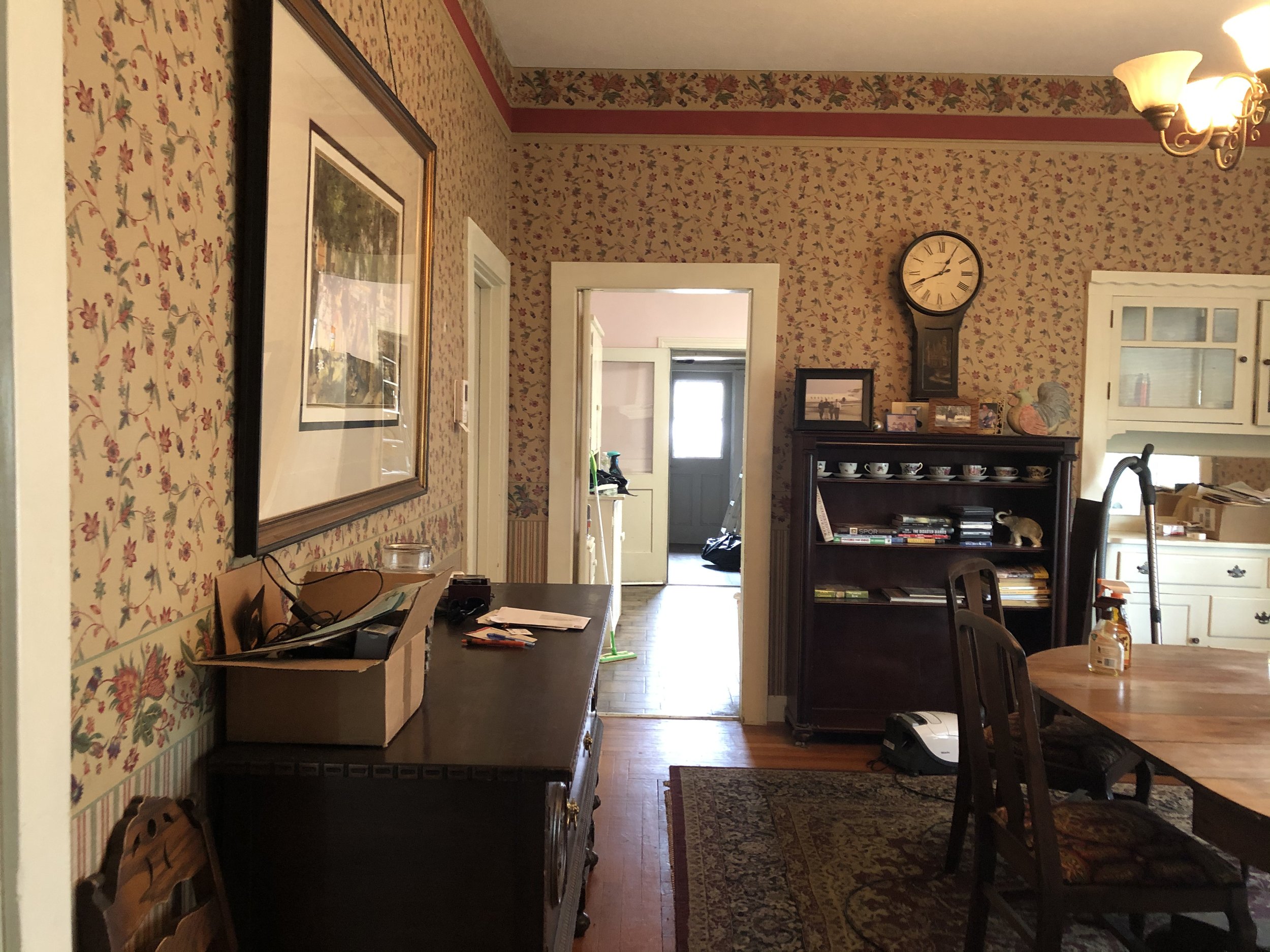 A cozy vintage-style kitchen with floral wallpaper, a wooden dining table, and a dark wood hutch displaying decorative teacups and framed photos, illuminated by a ceiling sconce and natural light from an open door.