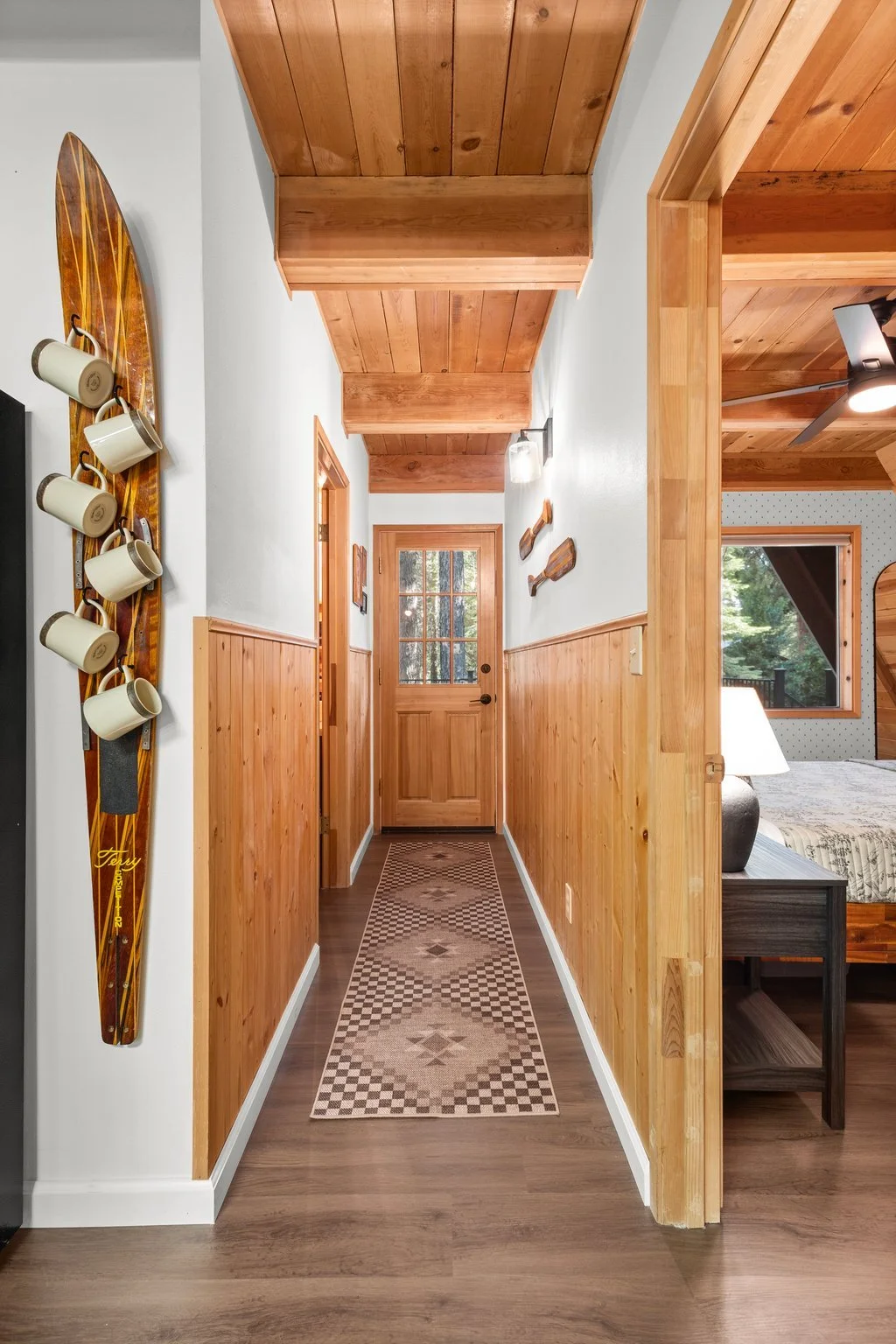 Wood-paneled hallway with a front door at the end, decorated with wall-mounted oars, and a woven rug on the floor leading to the door. A bedroom is visible to the right.