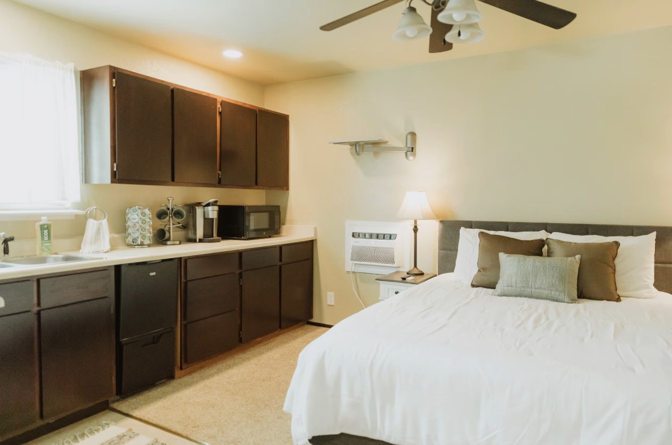 A bedroom with a large bed, brown and green pillows, a nightstand with a lamp, a window with white curtains, and a kitchenette with dark wood cabinets, a microwave, and coffee-making equipment.
