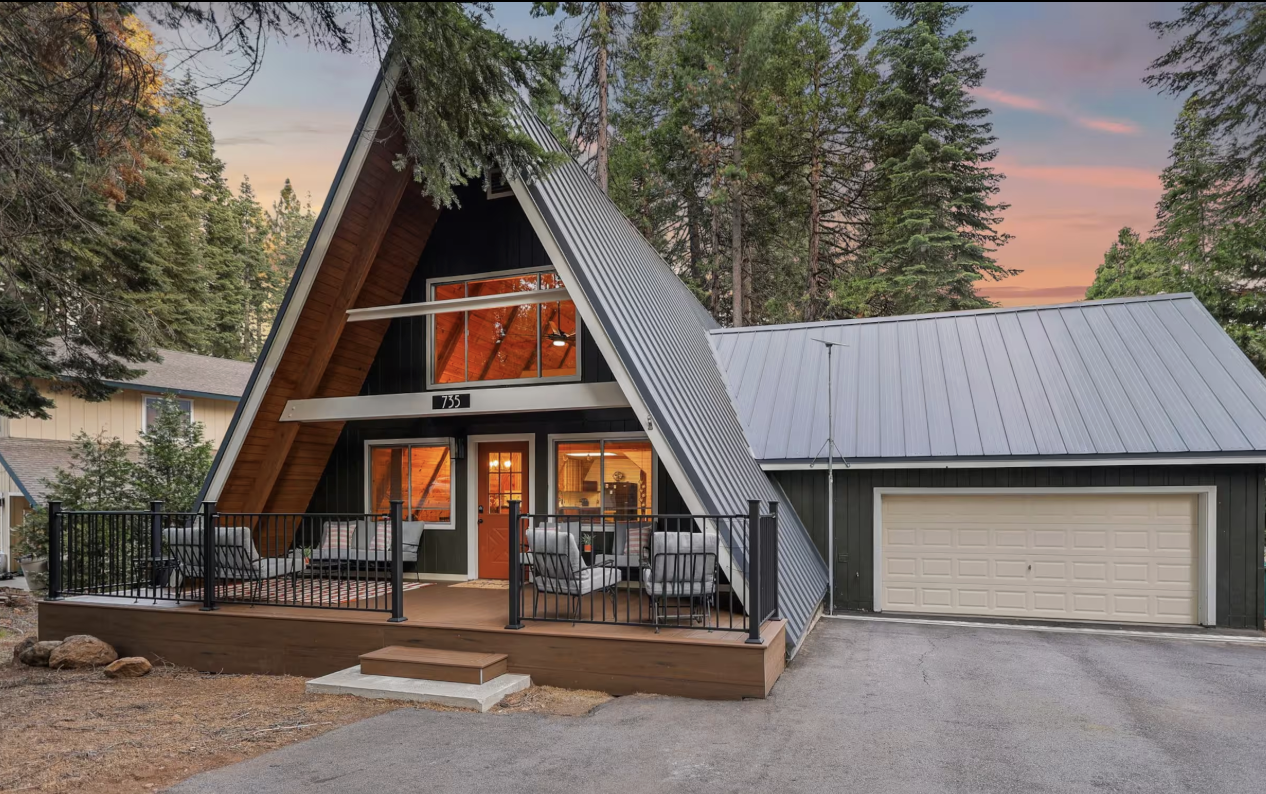 Modern A-frame house with a black exterior, large front porch with outdoor seating, overlooking a wooded yard, at sunset.