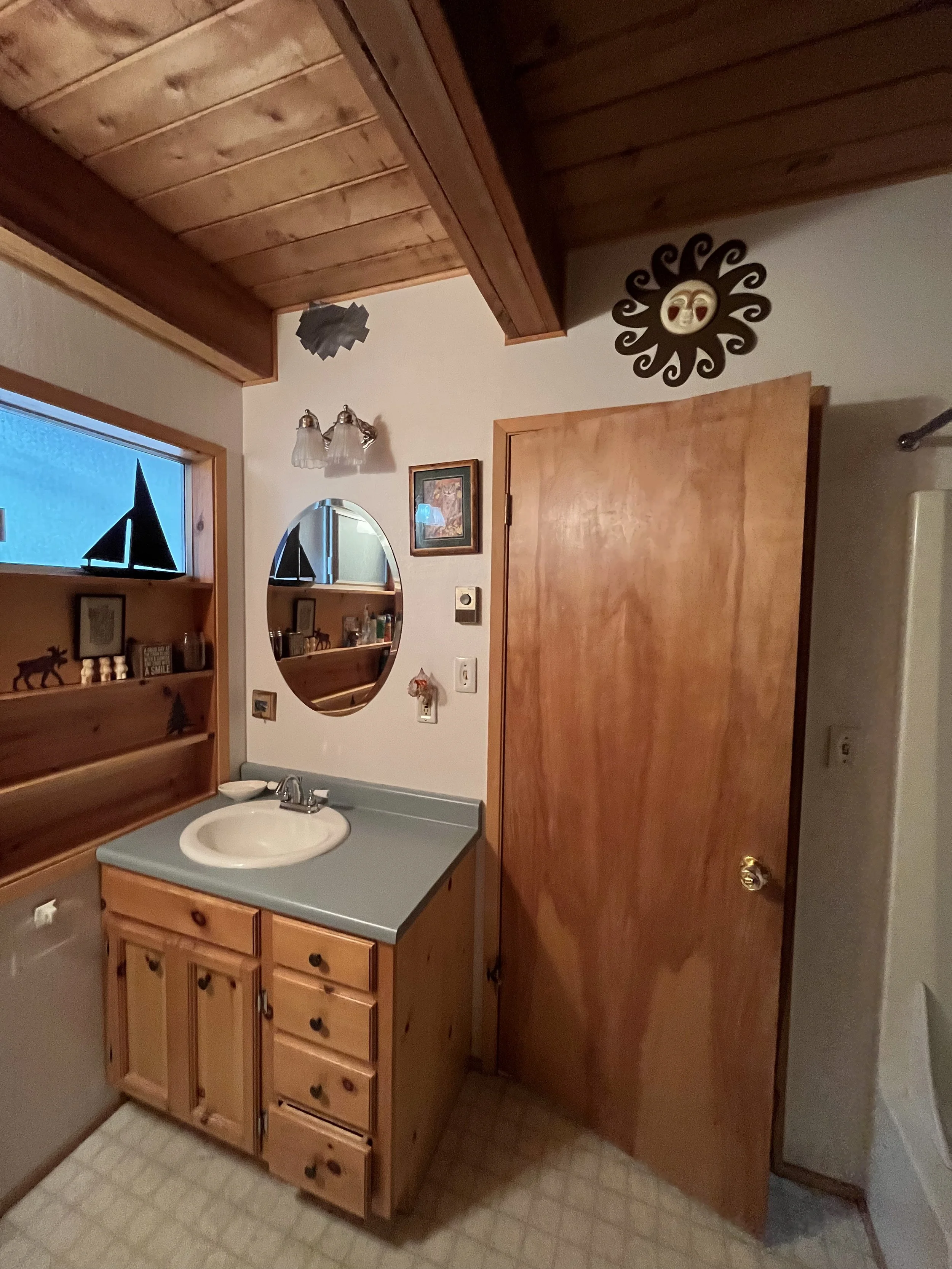 A bathroom with wooden cabinets, a small sink, a mirror, and a decorative black sun with a face on the wall.