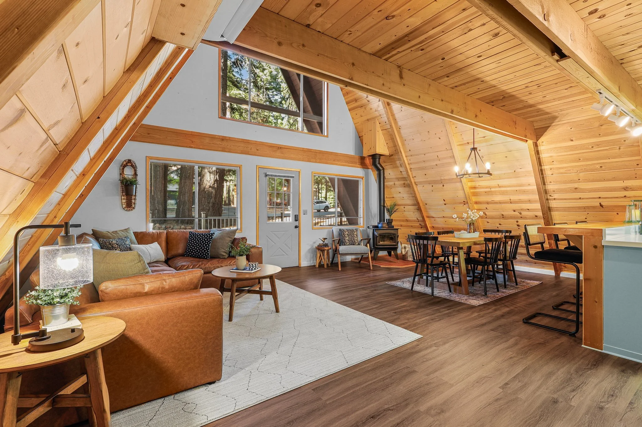 Interior of a cozy living space with wood-paneled A-frame ceiling, large windows showing outdoor trees, a brown leather sofa with decorative pillows, a small side table with a plant, a dining area with a wooden table and black chairs, and warm lighti