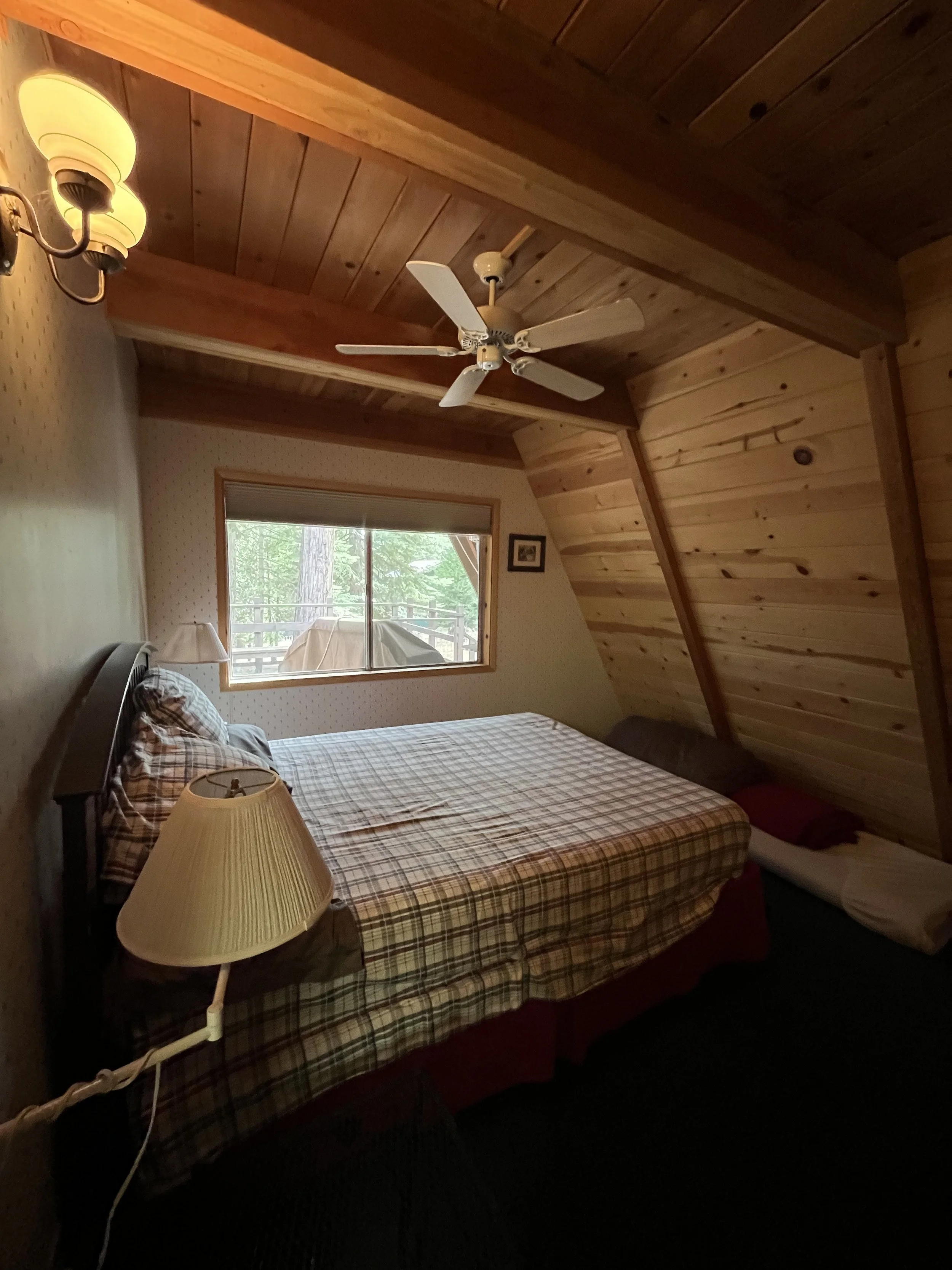 Inside a cozy bedroom with wooden walls and ceiling, a bed with a plaid bedspread, a nightstand with a lamp, a window showing green trees outside, and a ceiling fan.
