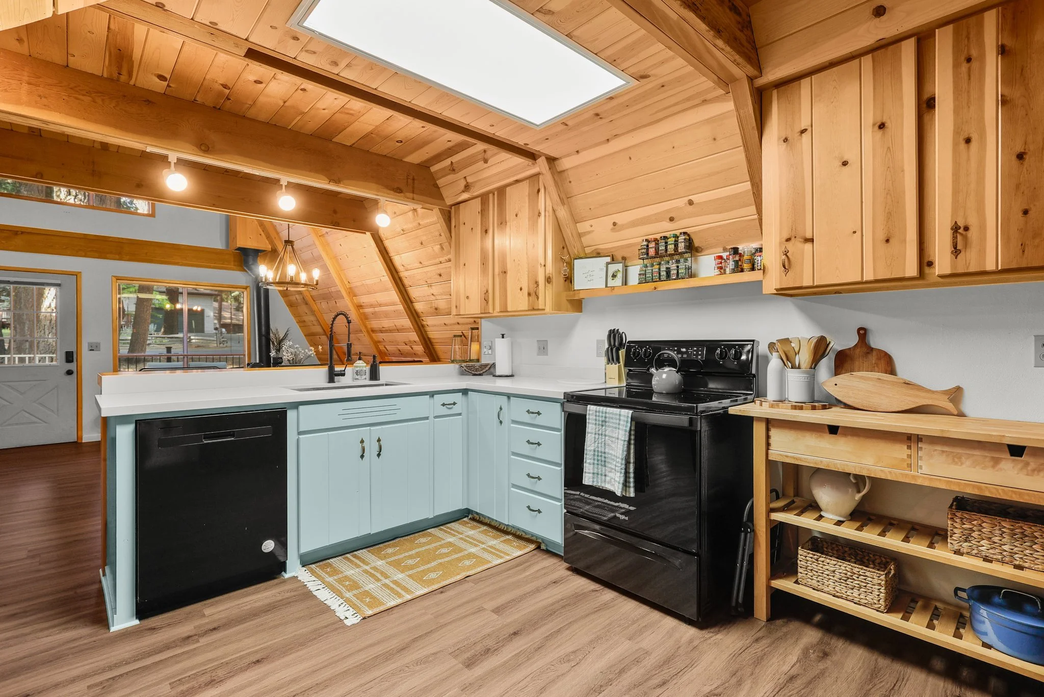 Inside view of a rustic kitchen with wooden ceiling and cabinets, white countertops, black appliances, and open shelving with baskets and jars.
