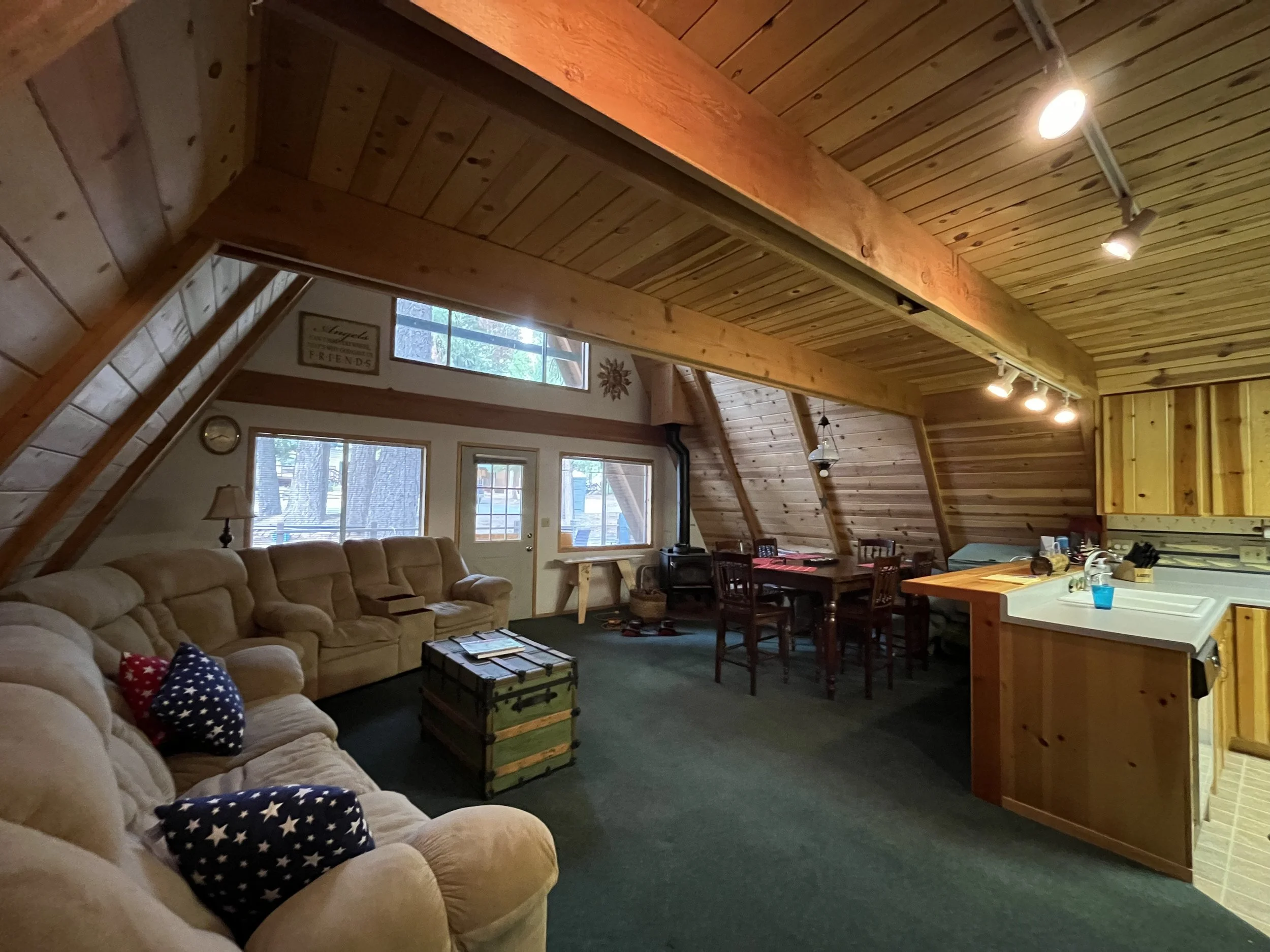 A cozy living room with wooden paneling on the walls and ceiling, featuring a beige sectional sofa with patriotic star-printed pillows, a wooden chest used as a coffee table, large windows letting in natural light, and a kitchen area with a wooden is