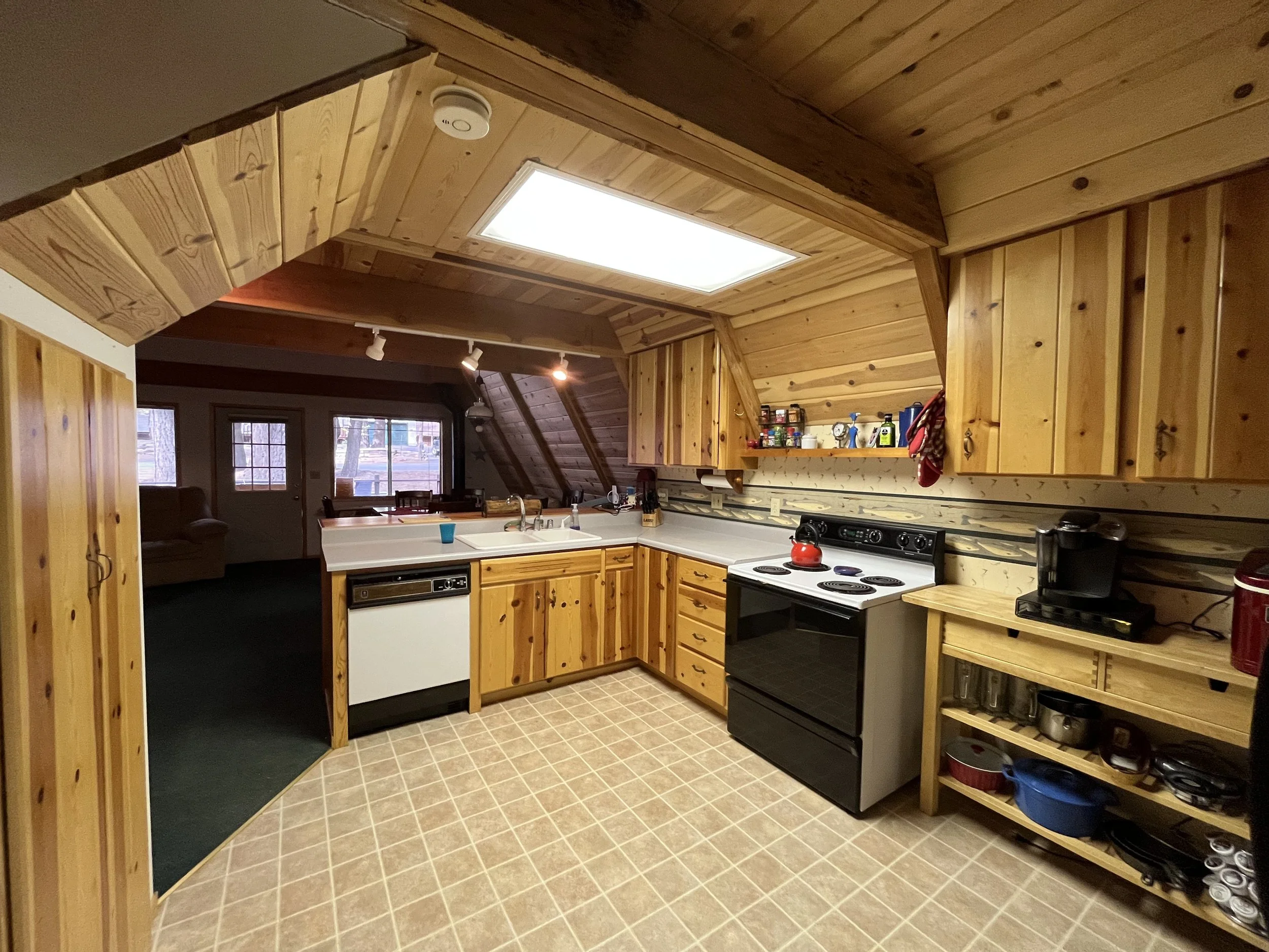 A cozy kitchen with wooden walls and cabinets, a tiled floor, a ceiling light, and a stove with a red kettle. There is a small open shelving unit with pots and dishes, and some cleaning supplies on the counter.