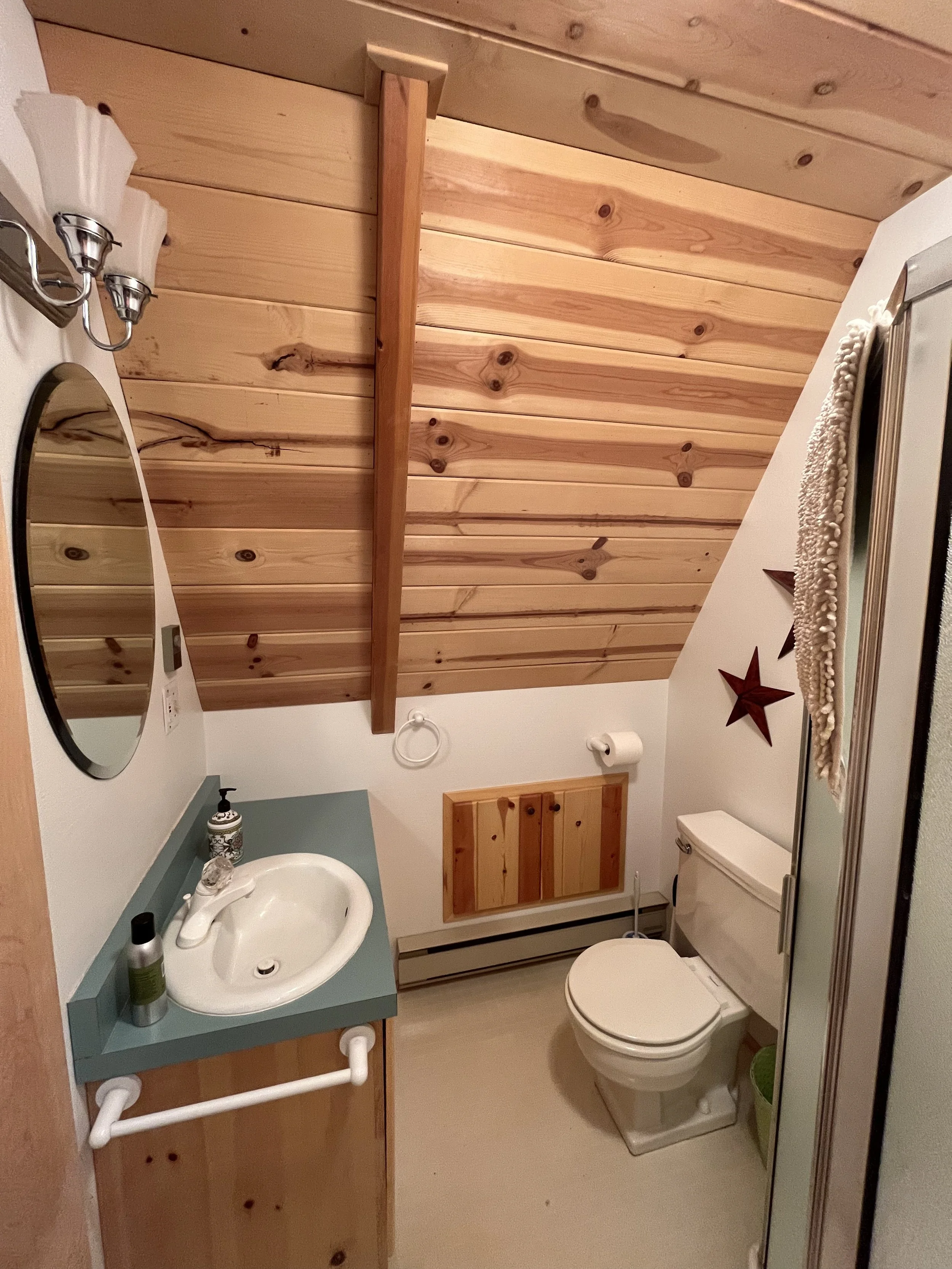 A small bathroom with wooden ceiling and wall, white toilet, white sink with green countertop, and decorative stars on the wall. There is a mirror and a towel ring beside the sink.