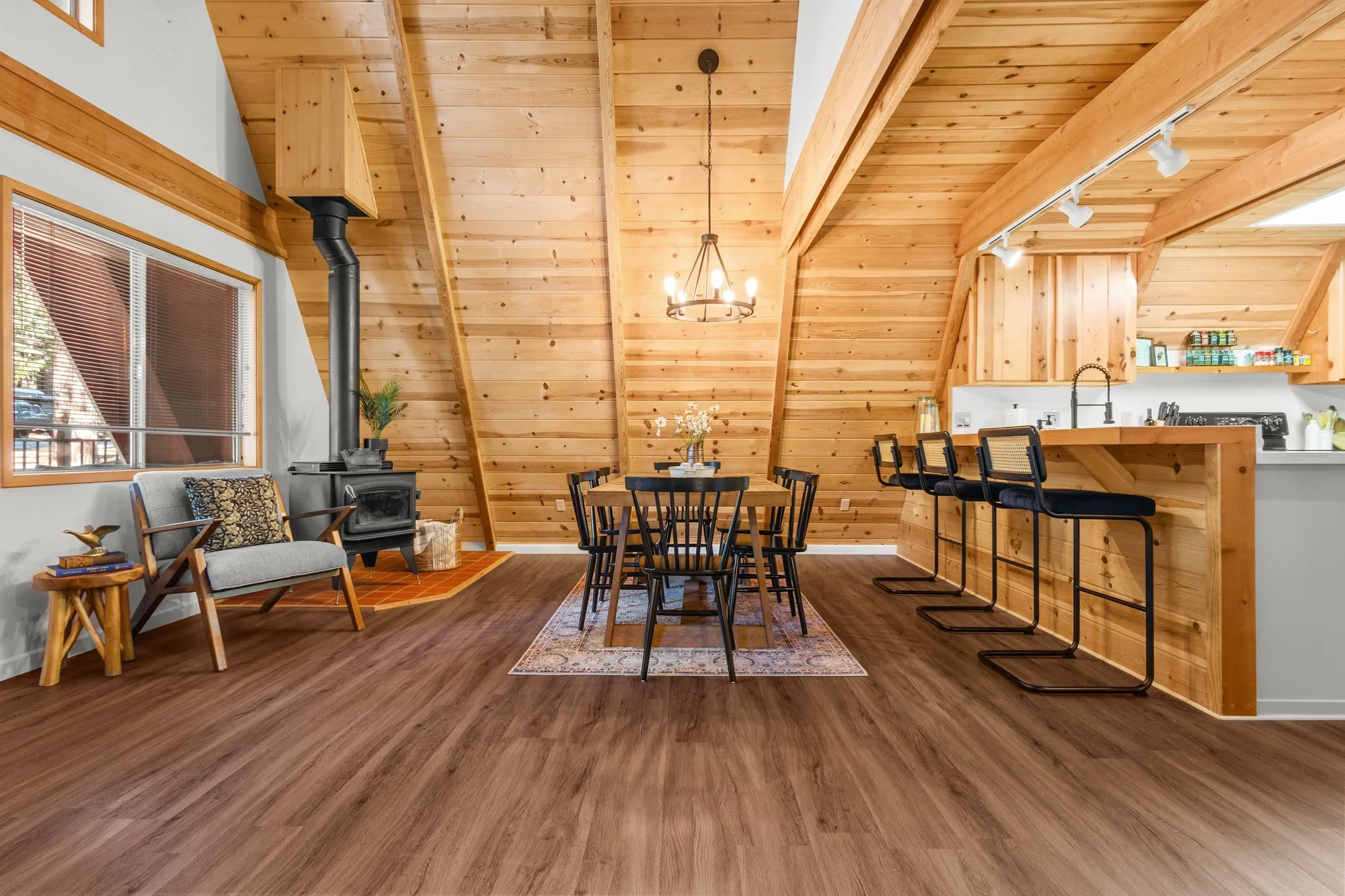 Interior of a wood-paneled living and kitchen area with a dining table, barstools, a sitting area with a chair and small table, and a wood stove, featuring vaulted ceilings and track lighting.