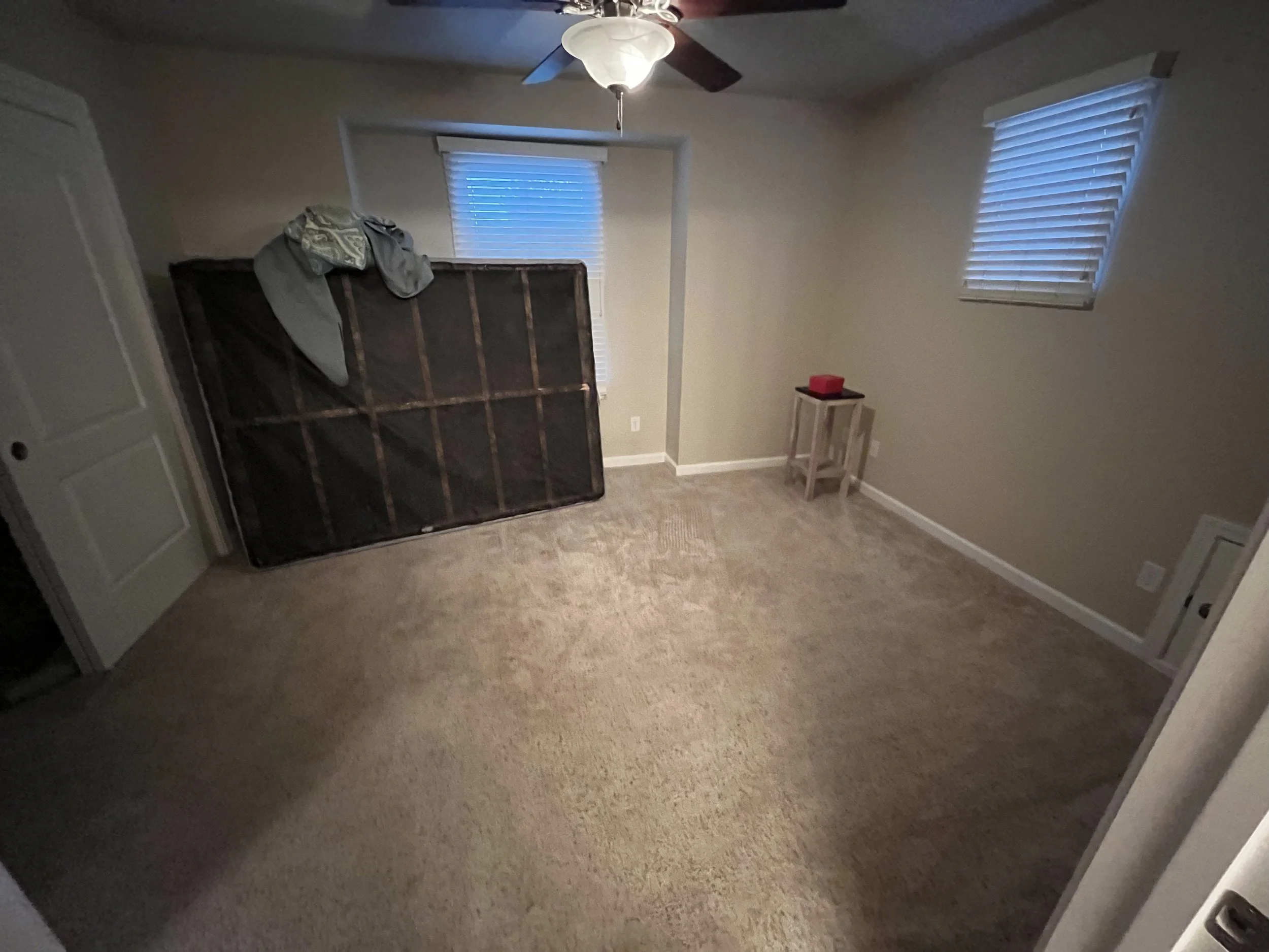 A room with beige walls and carpeted floor, containing a mattress leaning against the wall, a small wooden table with a red object on top, and two windows with blinds. A ceiling fan with a light fixture is on the ceiling.