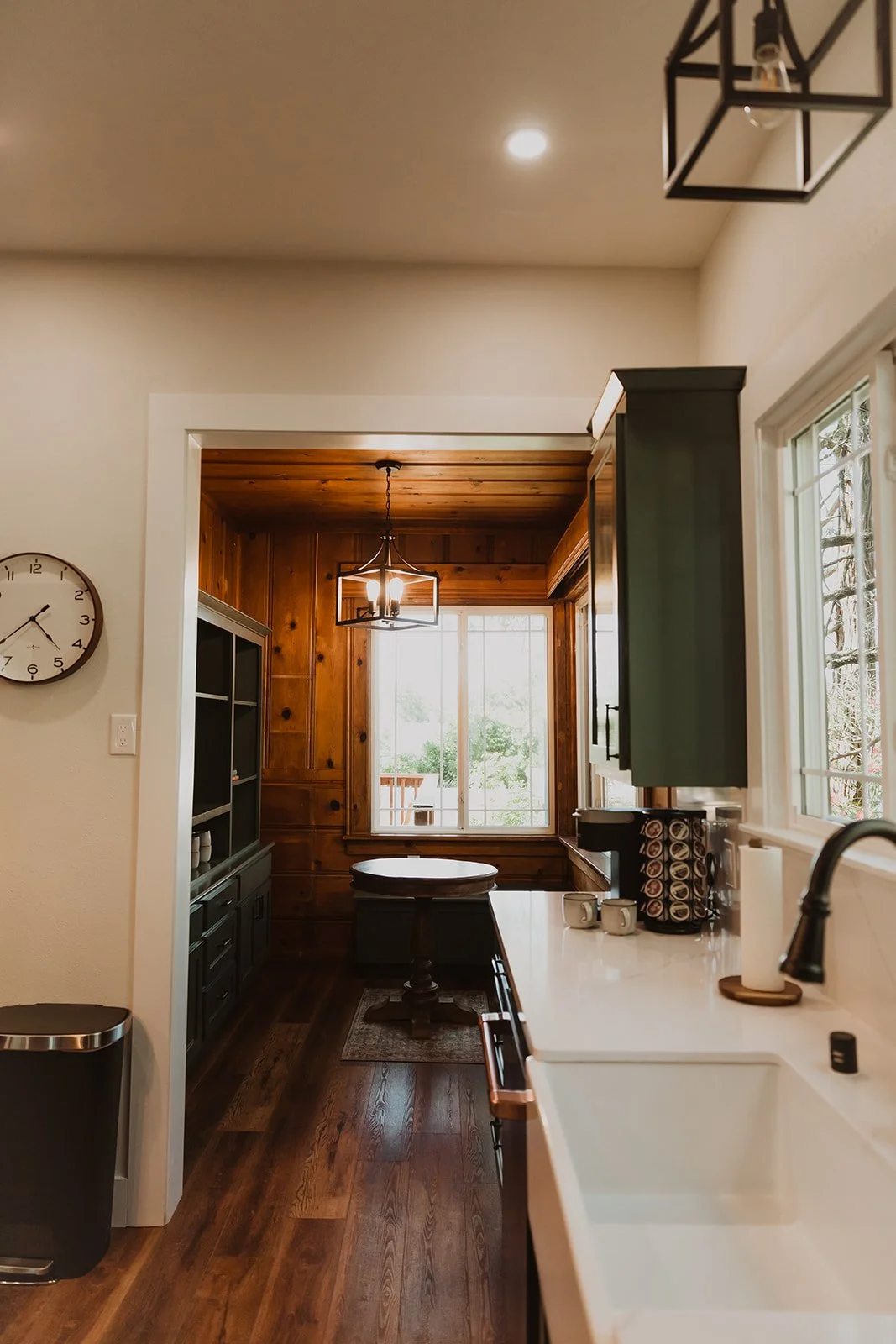 View of a kitchen with white countertops and a black faucet, green cabinets, a coffee station, and a window letting in natural light. A transition leads to a cozy nook with wood-paneled walls, a small round table, and a large window with trees outsid