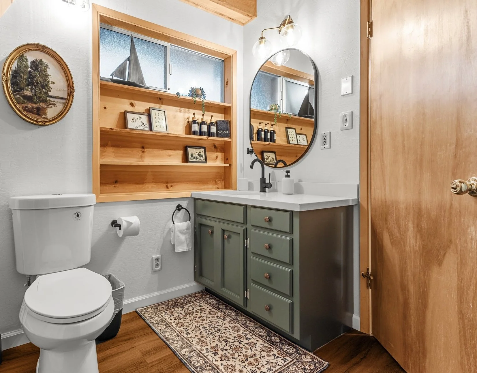 A bathroom with a white toilet, green vanity with drawer and cabinet doors, oval mirror, and wooden door. Wooden shelves hold framed pictures, soap dispensers, and a small plant, with a sailboat decoration on the frosted window.