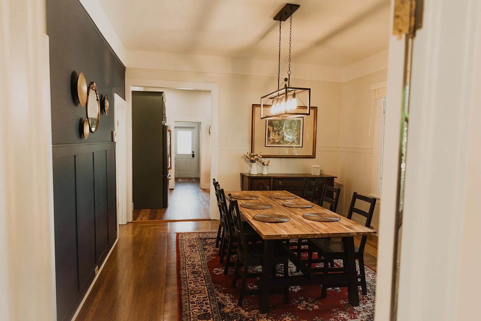 Dining room with wooden table, six black chairs, a patterned red area rug, black sideboard, two vases with flowers, framed artwork, and hanging modern light fixture, with door leading to foyer visible in background.