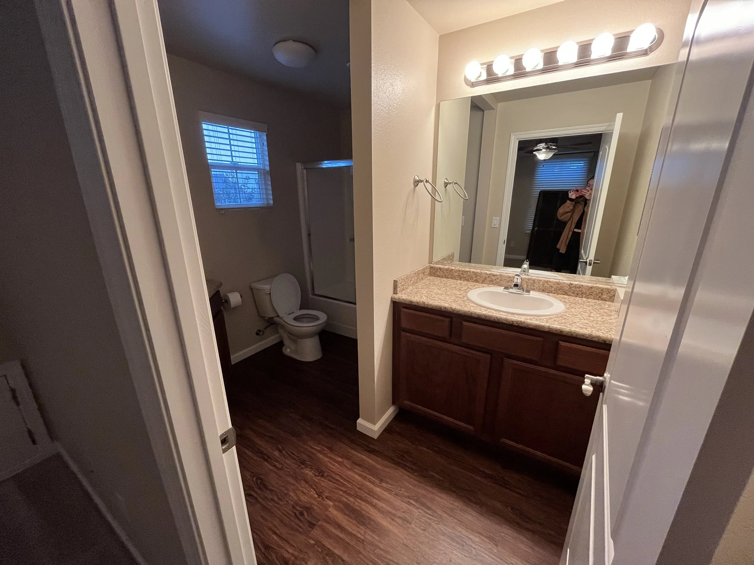 Bathroom with granite countertop, mirror, vanity lights, and a visible toilet in a separate area with a window showing a tree outside.