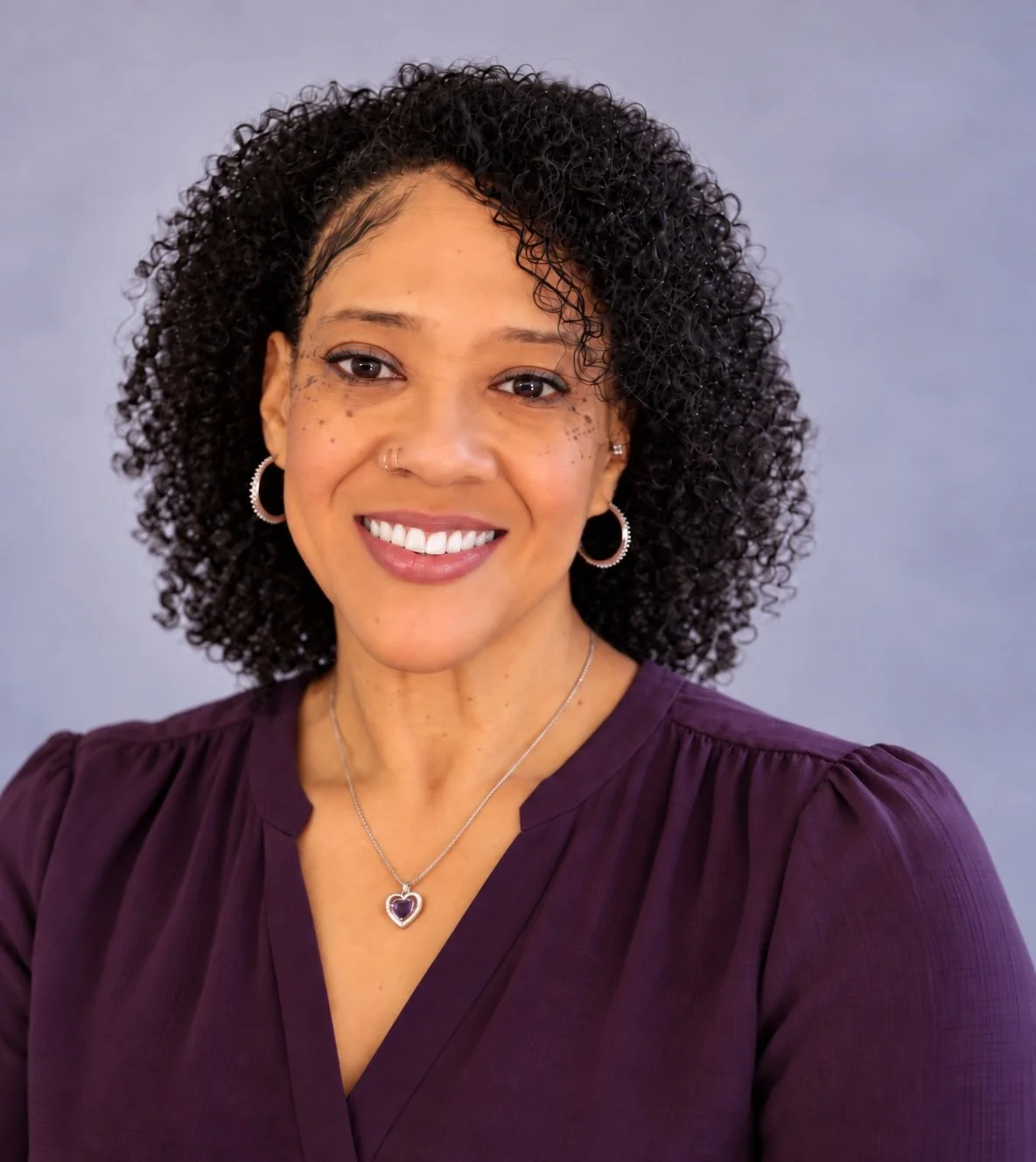 A woman with curly dark hair smiling at the camera, wearing a purple top, hoop earrings, and a heart-shaped necklace.
