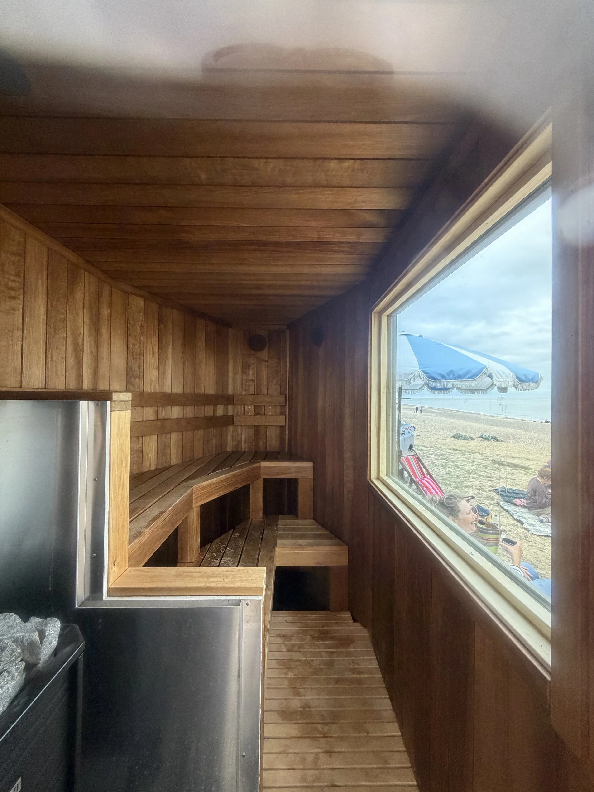 Wood-paneled interior of a beachside hut or cabana with a large window showing people relaxing outside on a sandy beach under umbrellas.