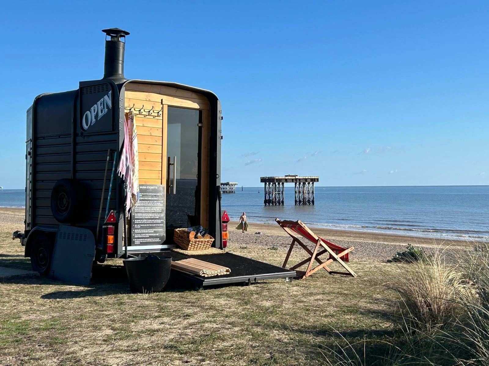 A black horsebox that has been converted into a sauna set up on a beach with a deck chair, beach towel, and a person walking along the shoreline, with pier structures in the ocean under a clear blue sky.