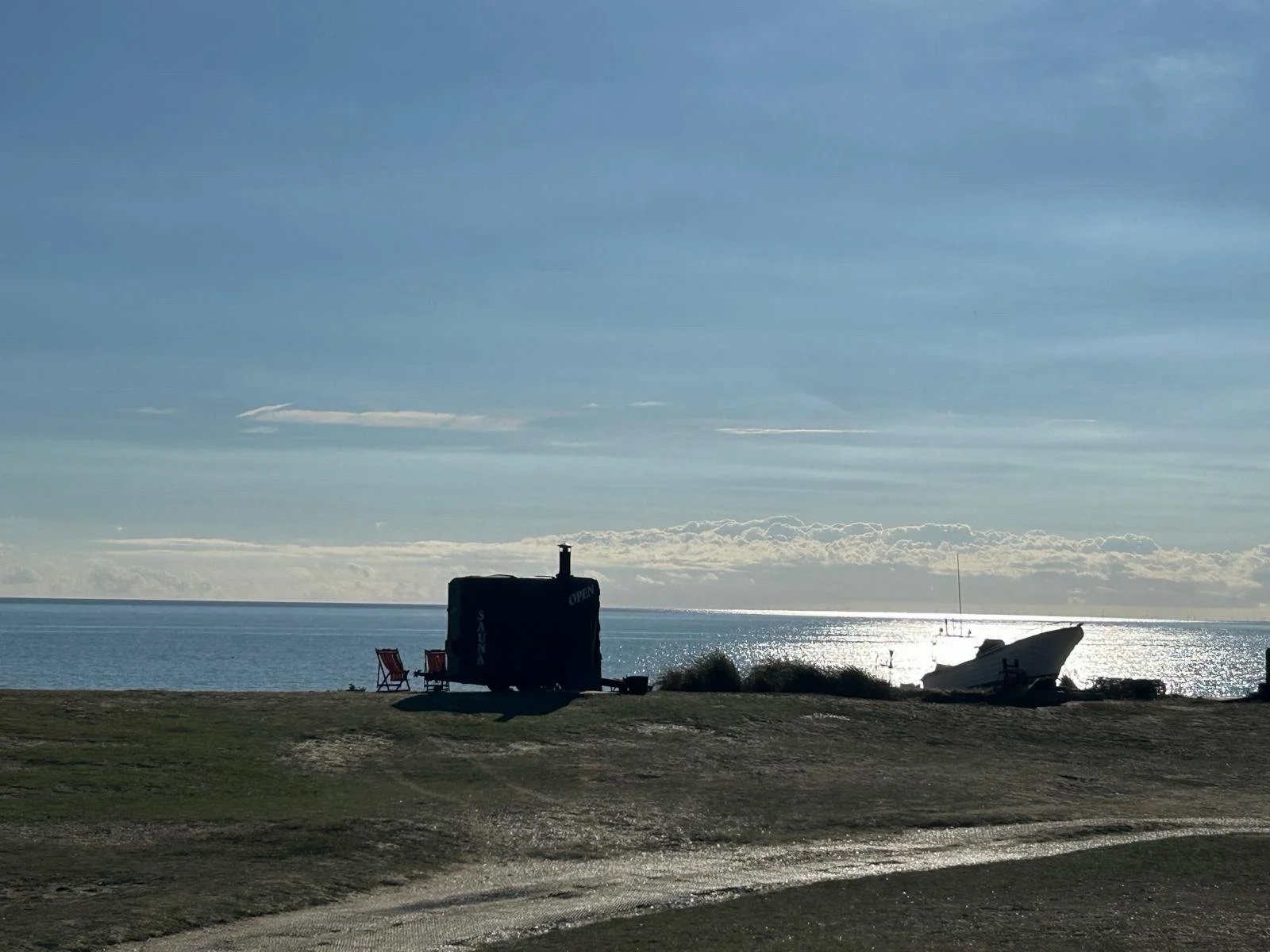 A peaceful beach scene with a boat stranded on the shore, a small shelter with a sign that says 'Open', and two chairs near the water. The sky is partly cloudy with the sun reflecting on the ocean.