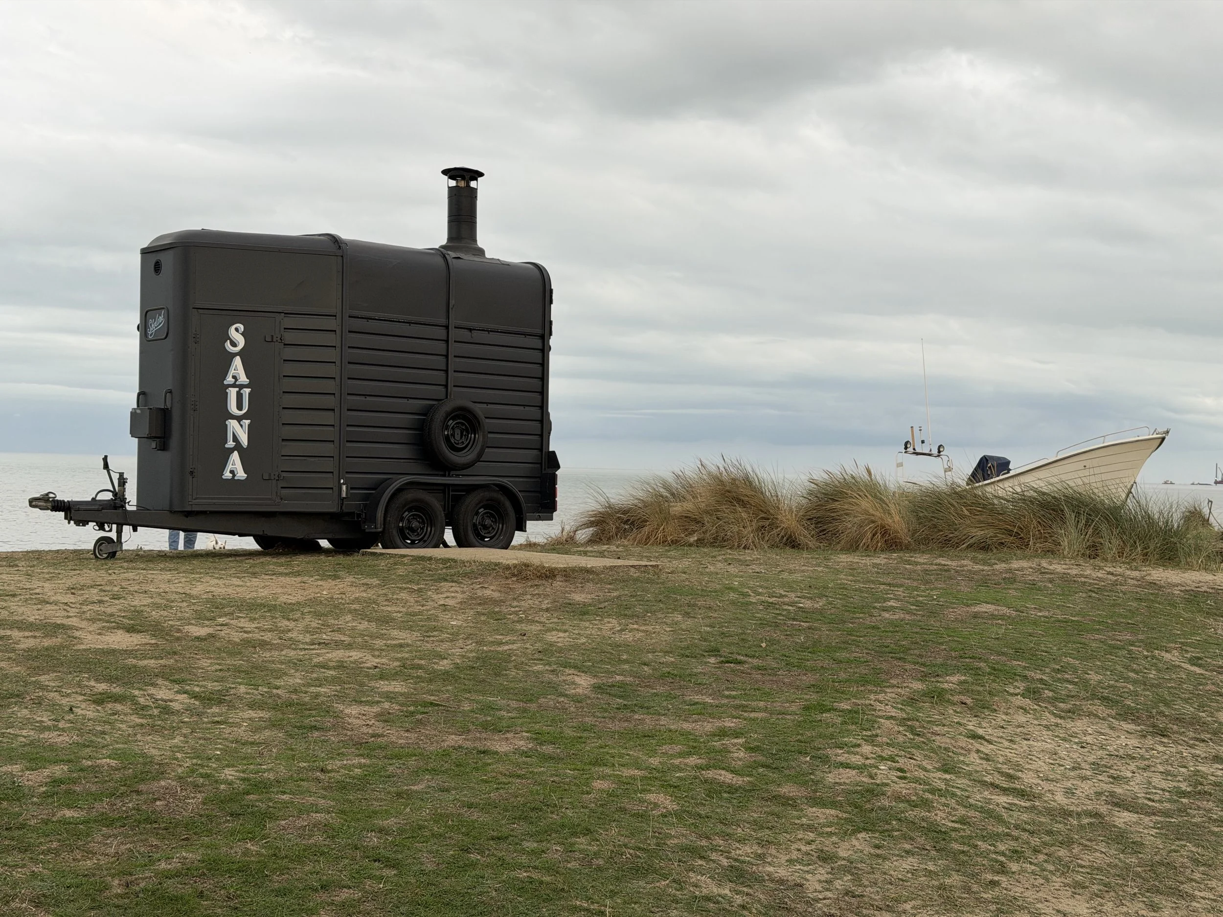 A black food truck named 'Sauna' with a chimney on top, parked on a grassy area near the beach under cloudy skies. There is a boat and some equipment visible in the background.