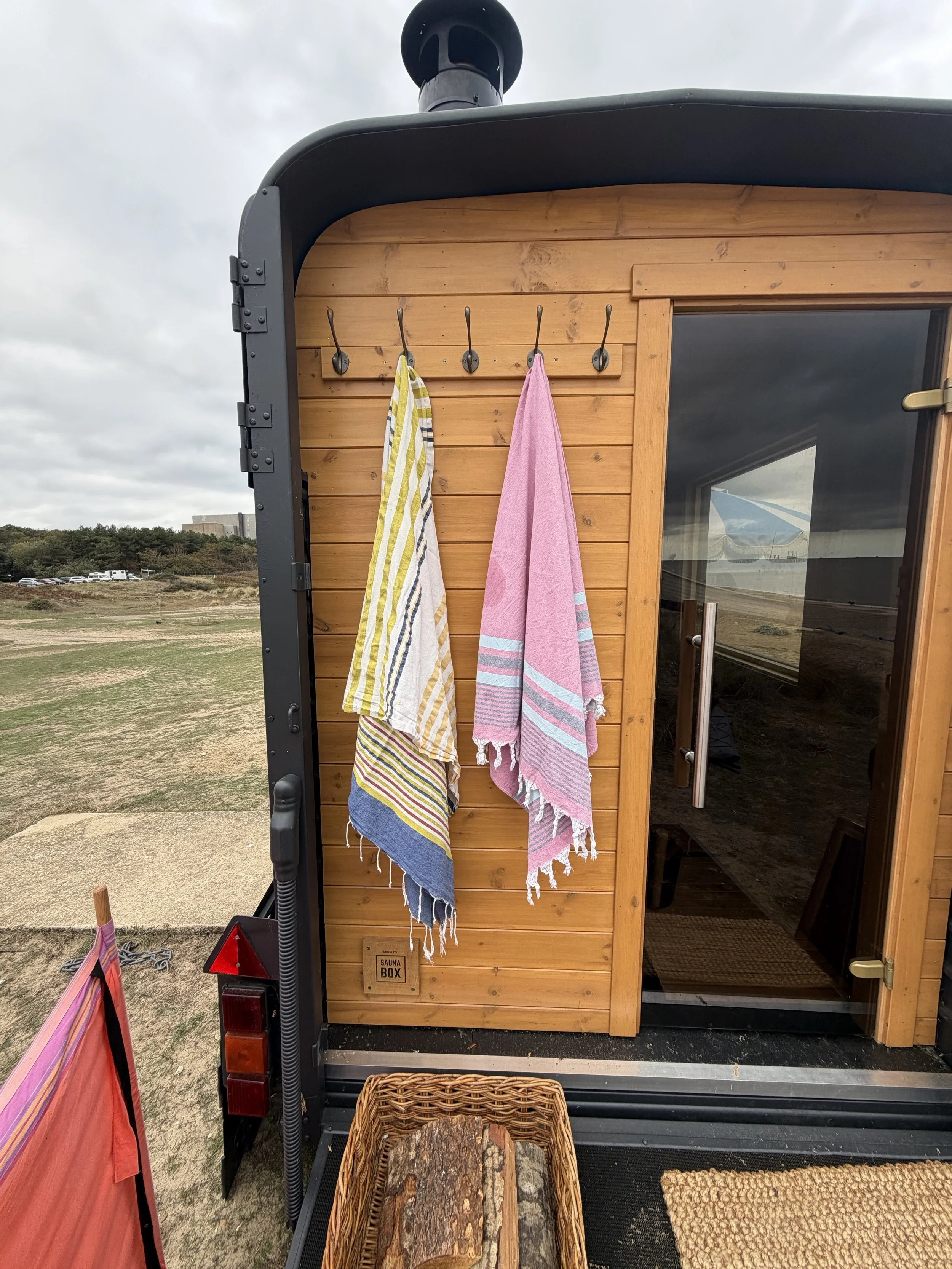 Small wooden sauna with hooks holding colorful towels, a sign indicating 'Sauna Box,' a woven basket with firewood in front, set outdoors on a cloudy day.