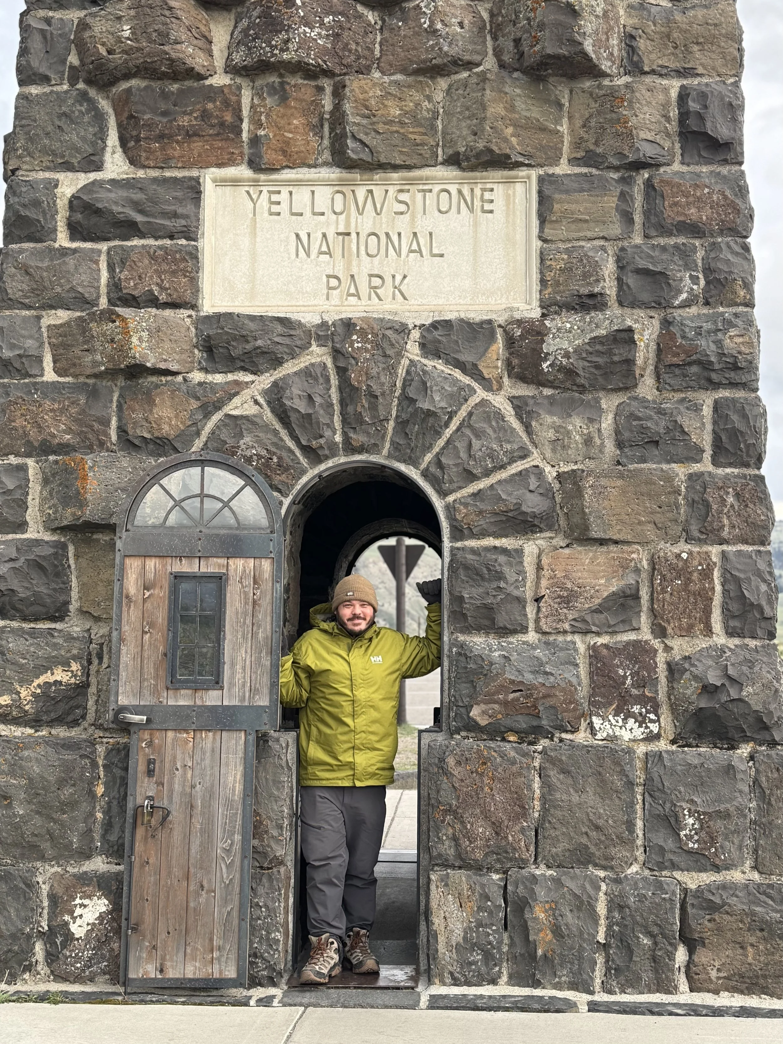 A person standing at the entrance of Yellowstone National Park behind a stone archway, smiling, wearing a green jacket and a brown beanie.